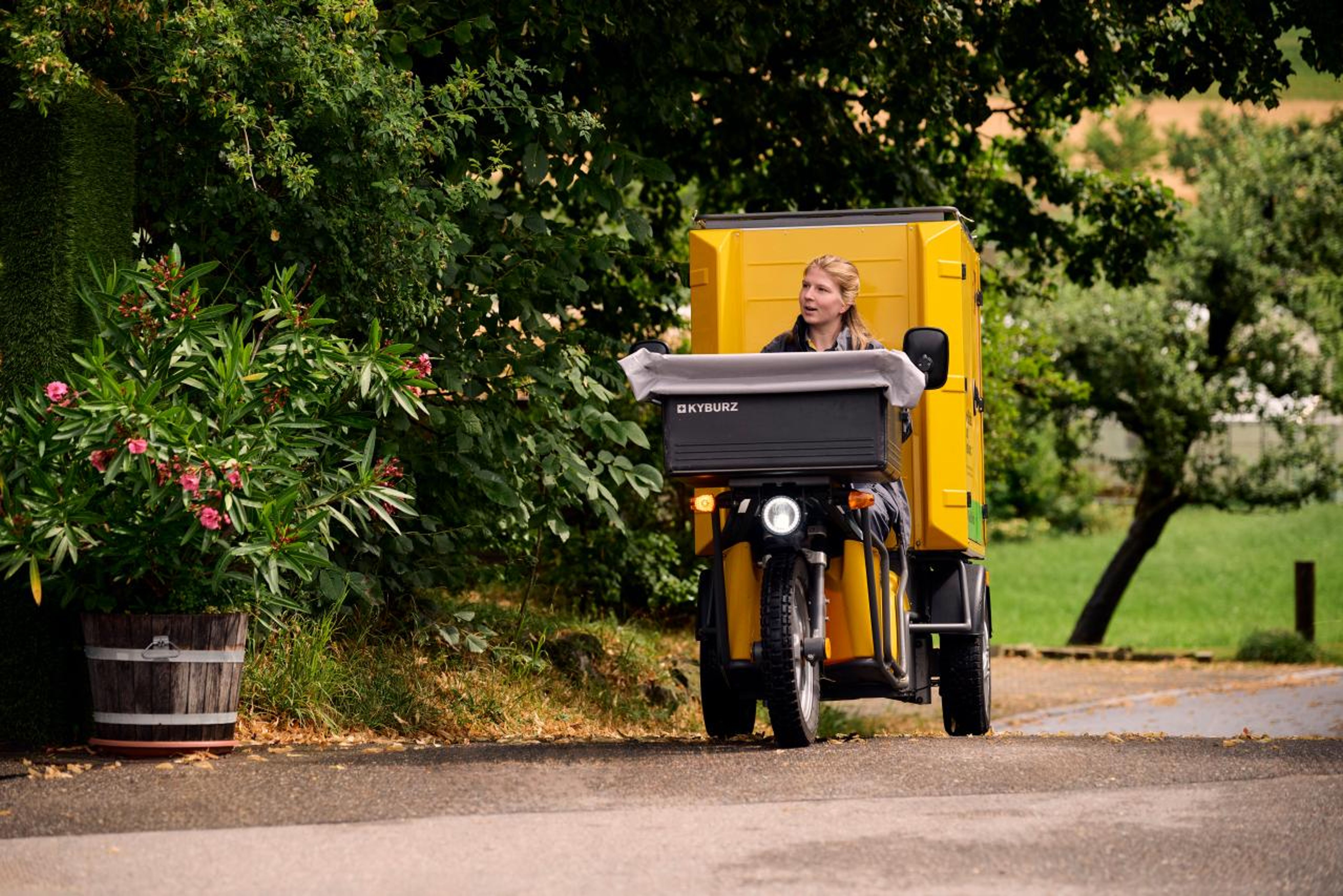 Une collaboratrice de la Poste conduit un véhicule électrique à trois roues de la marque Kyburz le long d’une route rurale. Le véhicule est jaune et équipé d’un local de livraison de la Poste. Les environs sont verdoyants et pittoresques, avec des arbres et des pots de fleurs. L’atmosphère est calme et idyllique,