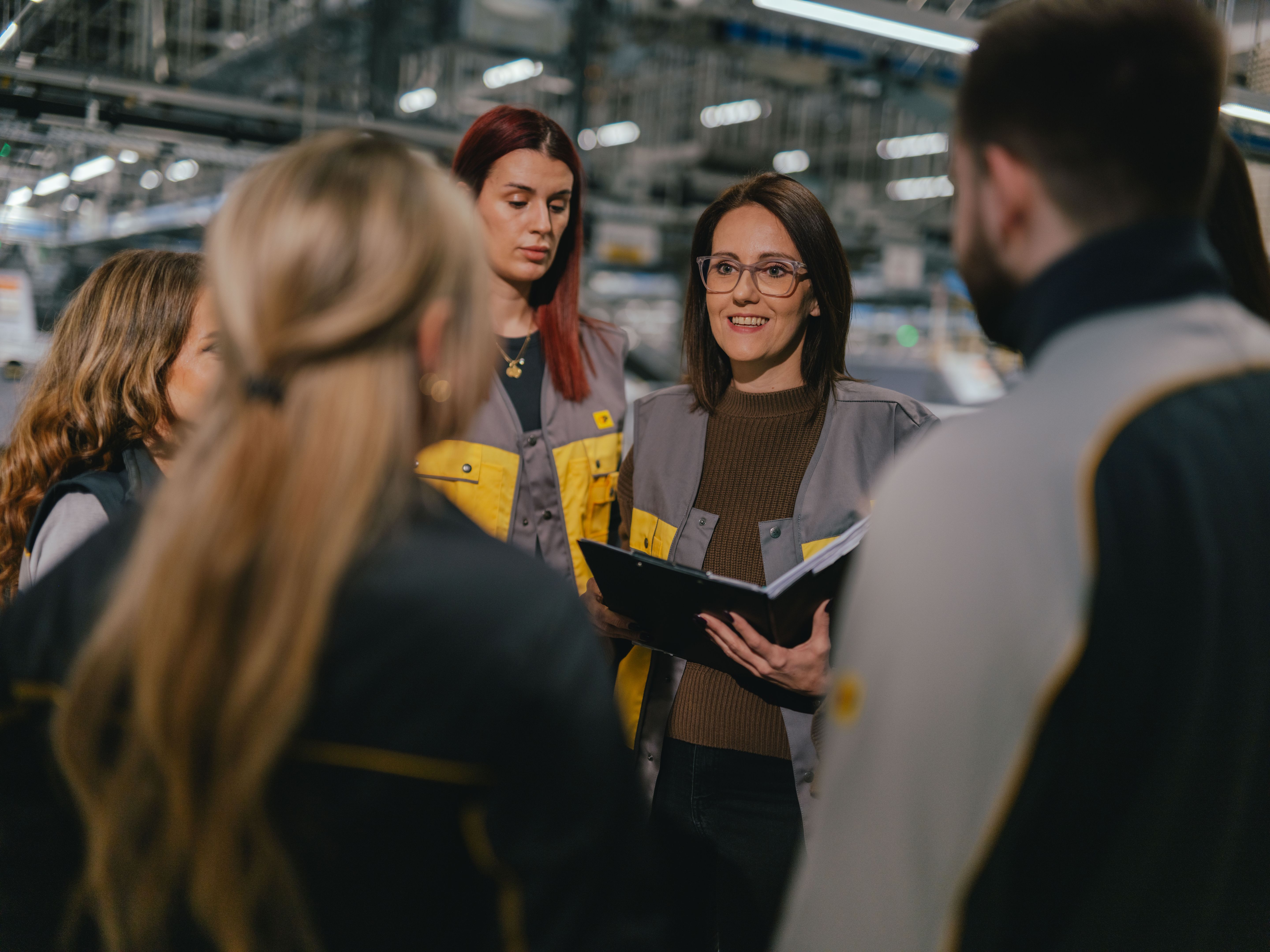 Un groupe de personnes se tient ensemble dans un hall de production ou de logistique. Plusieurs personnes portent des vêtements de travail gris et jaunes. Une personne tient un classeur ouvert, tandis que le groupe forme un cercle. À l’arrière‑plan, on aperçoit des machines, des étagères et un éclairage de plafond lumineux.