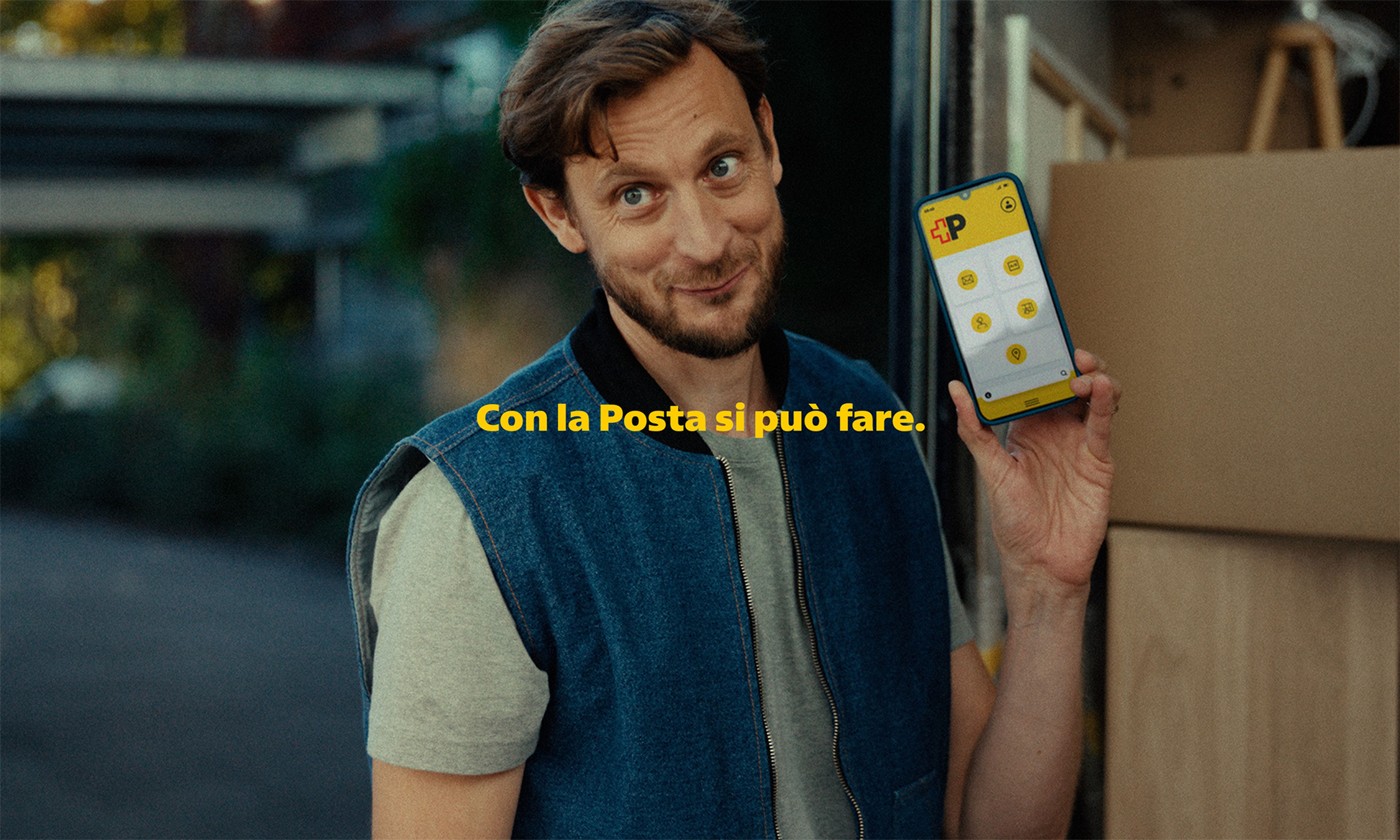 Una persona si trova davanti a un veicolo di trasporto aperto e tiene sollevato uno smartphone che mostra un’interfaccia gialla con diverse icone. Davanti alla persona ci sono scatole e superfici in legno. Sullo sfondo sono visibili alberi, un edificio e una struttura coperta. Al centro dell’immagine compare il testo giallo: “Con la Posta si puo fare”