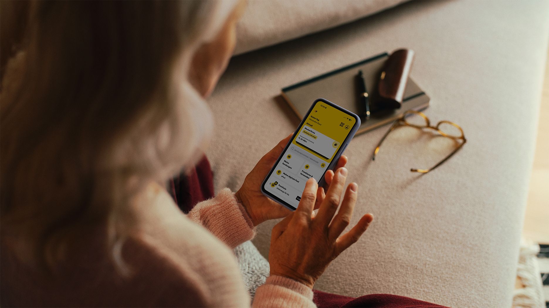Eine Frau sitzt auf einem Bett und bedient am Handy die Post-App.