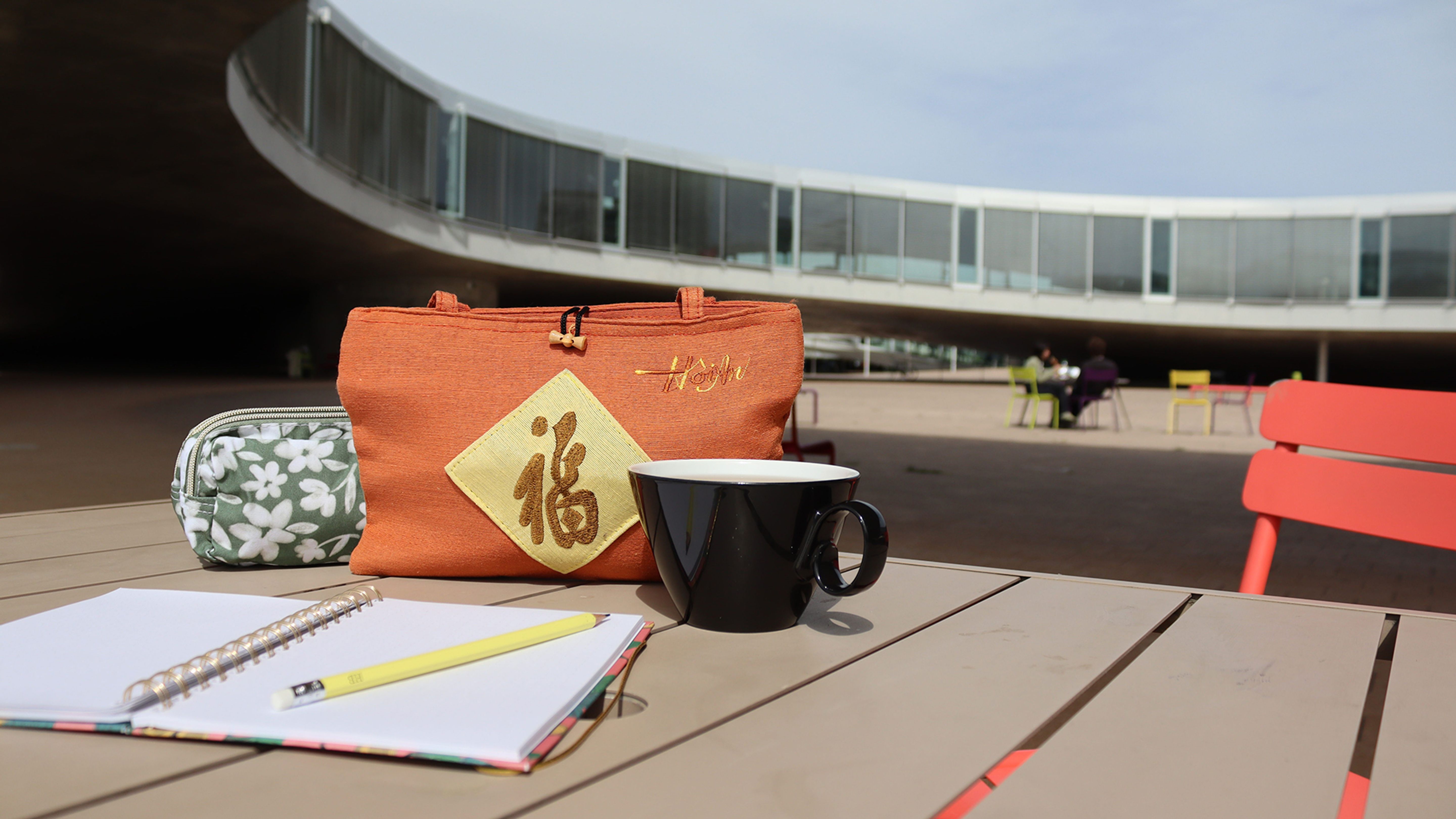 Au premier plan se trouvent un bloc-notes, un stylo jaune, une tasse noire et deux sacs sur une table devant une architecture moderne.