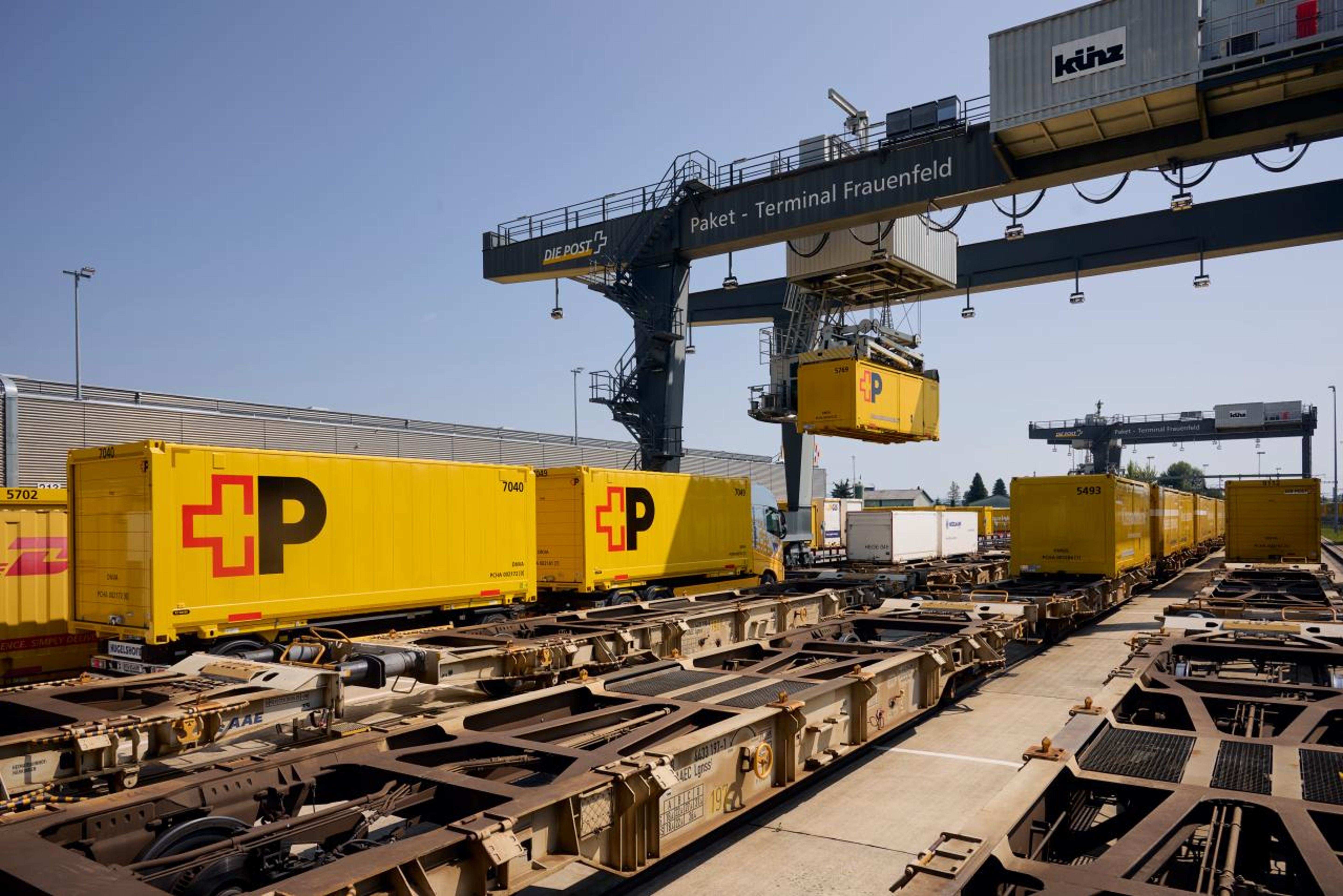 Yellow Swiss Post containers with the Swiss Post logo stand on railway carriages at the parcel terminal in Frauenfeld. A large crane lifts a container. The scene shows efficient logistics under a clear sky in a modern transhipment center. The atmosphere is bustling and structured.