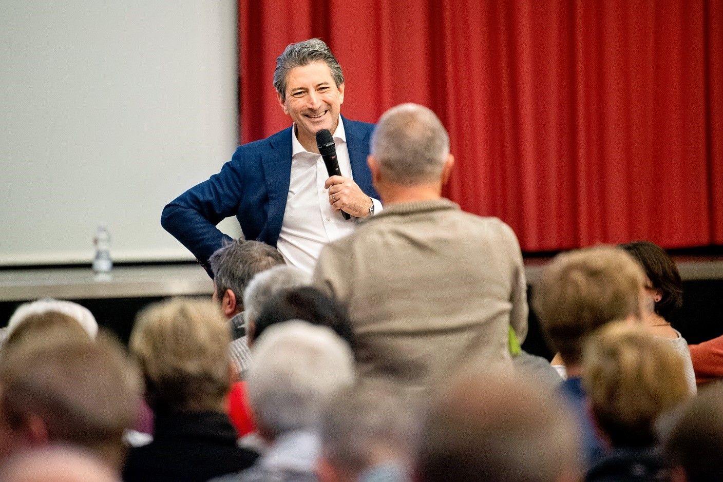 Ein Mann spricht in ein Mikrofon vor einem Publikum mit rotem Vorhang im Hintergrund.