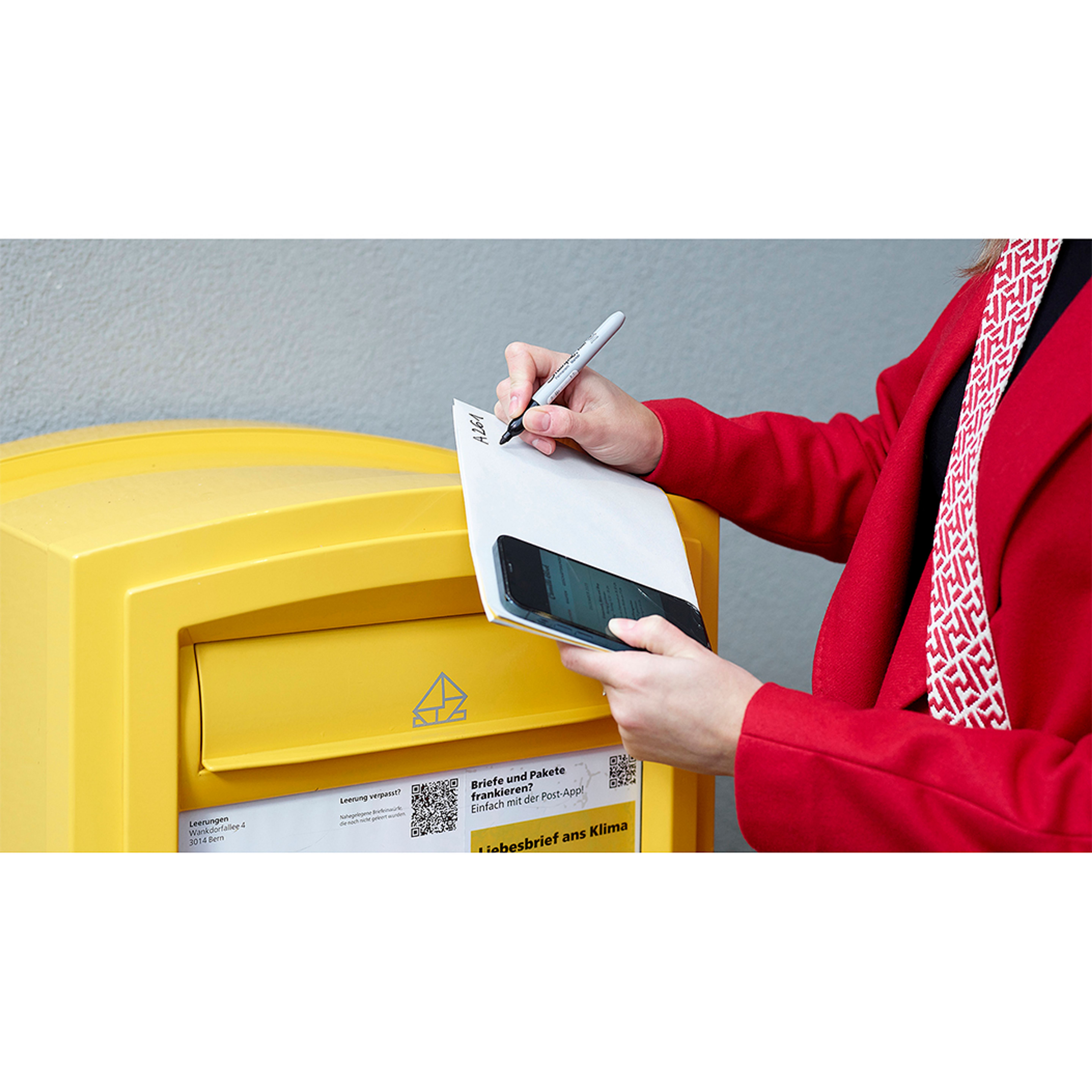 Eine Person schreibt auf einen Umschlag vor einem gelben Briefkasten mit einem Smartphone in der Hand.