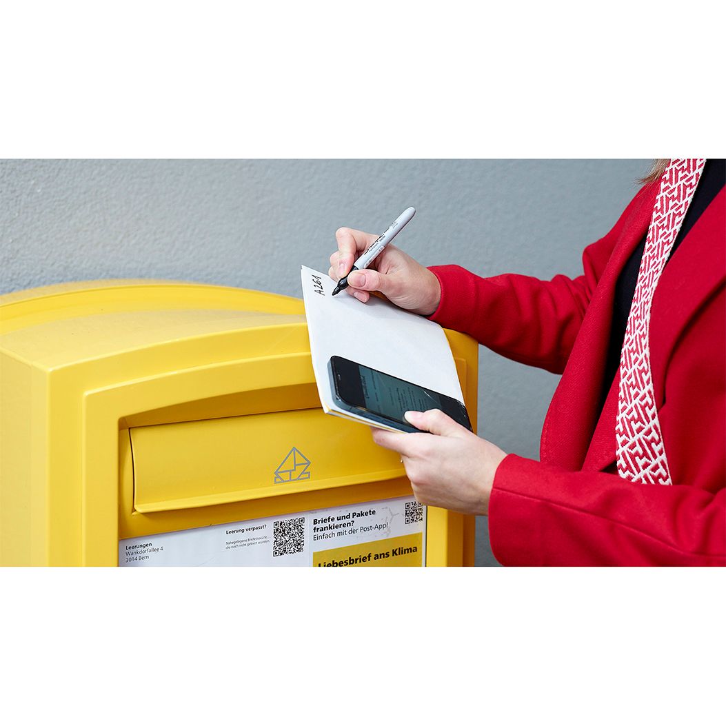 Eine Person schreibt auf einen Umschlag vor einem gelben Briefkasten mit einem Smartphone in der Hand.