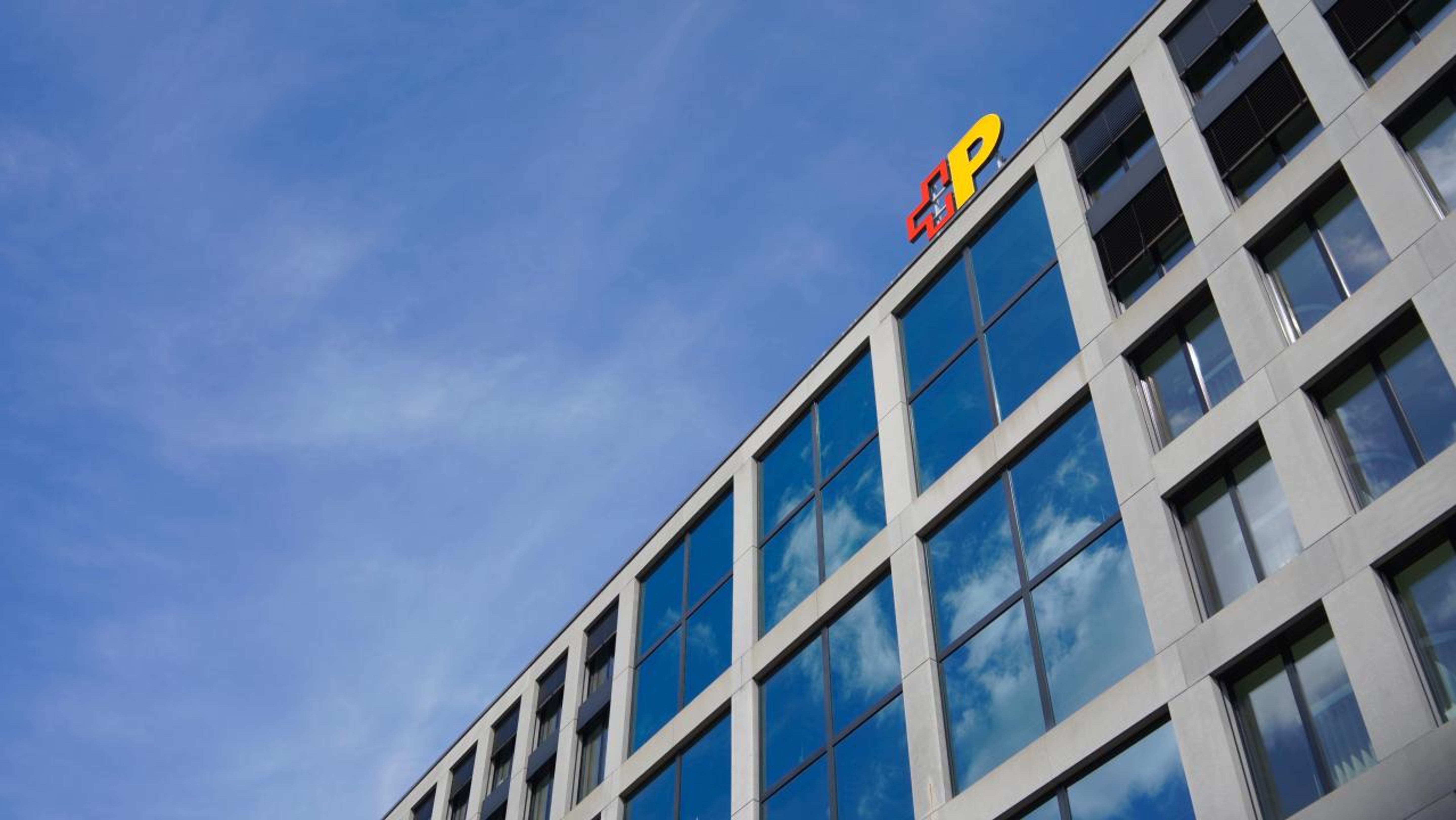 La photo montre la façade d’un immeuble de bureaux moderne avec de grandes vitres et un logo de la Poste sur le toit. Le ciel est clair et bleu, ce qui souligne l’architecture du bâtiment,