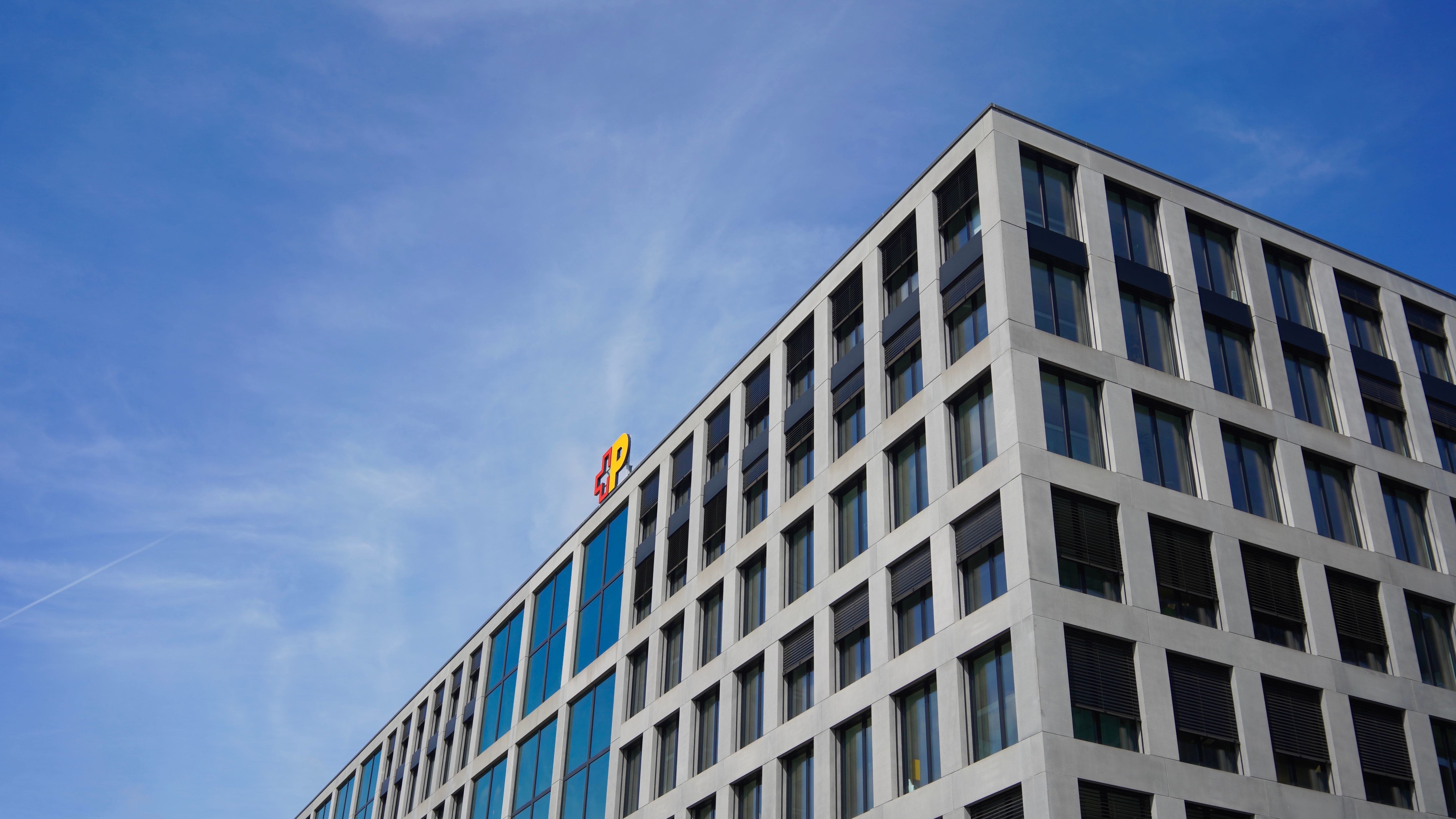 Das Bild zeigt die obere Ecke eines modernen Bürogebäudes mit vielen grossen Fenstern und einer Fassade aus Beton und Glas. Auf dem Dach ist das farbige Logo der Schweizerischen Post sichtbar. Der Himmel ist klar und blau, was eine helle und freundliche Atmosphäre schafft.