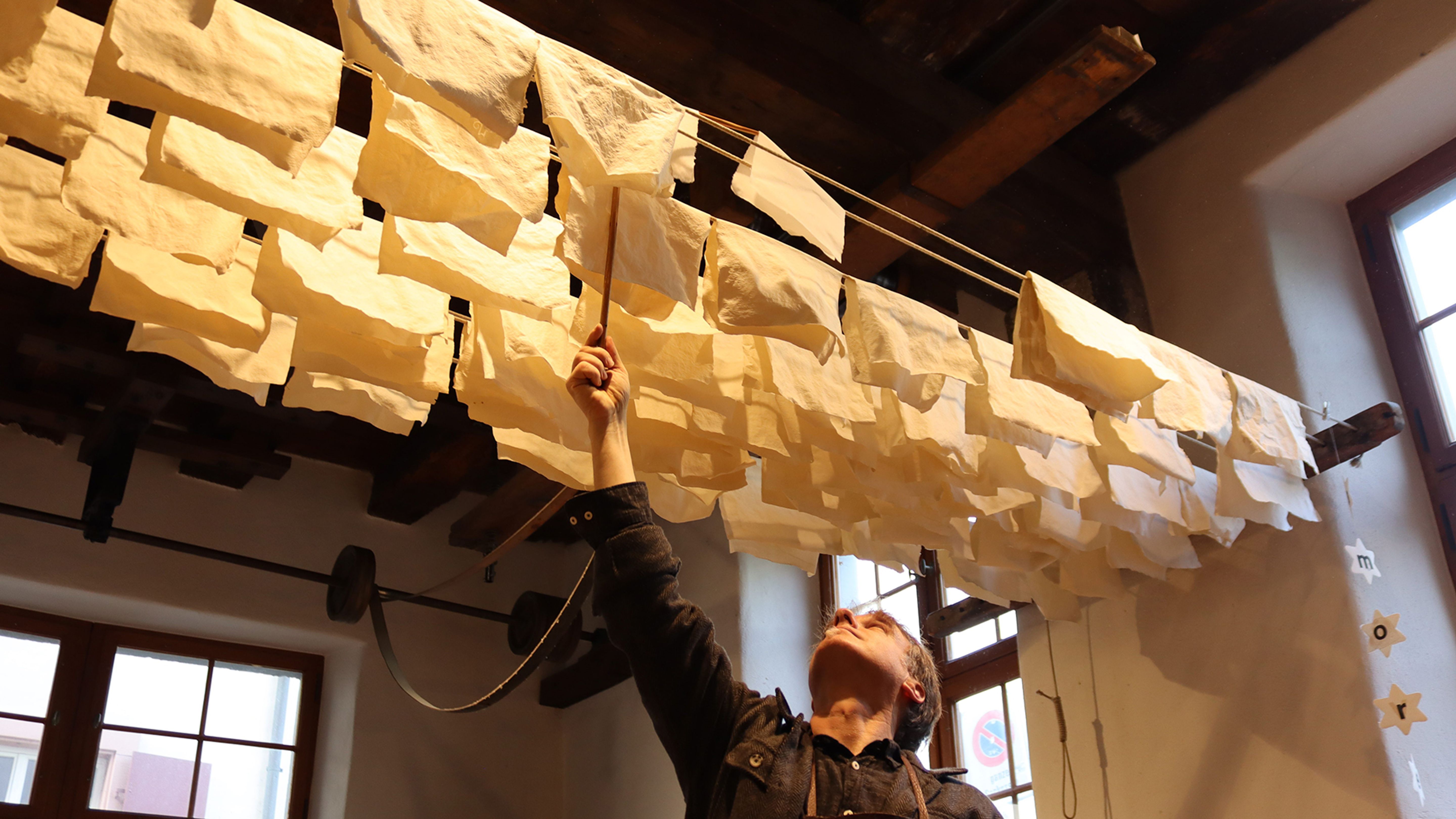 Eine Person inspiziert hängende gelbe Papierstücke in einem warm beleuchteten Raum mit Holzbalkendecke.