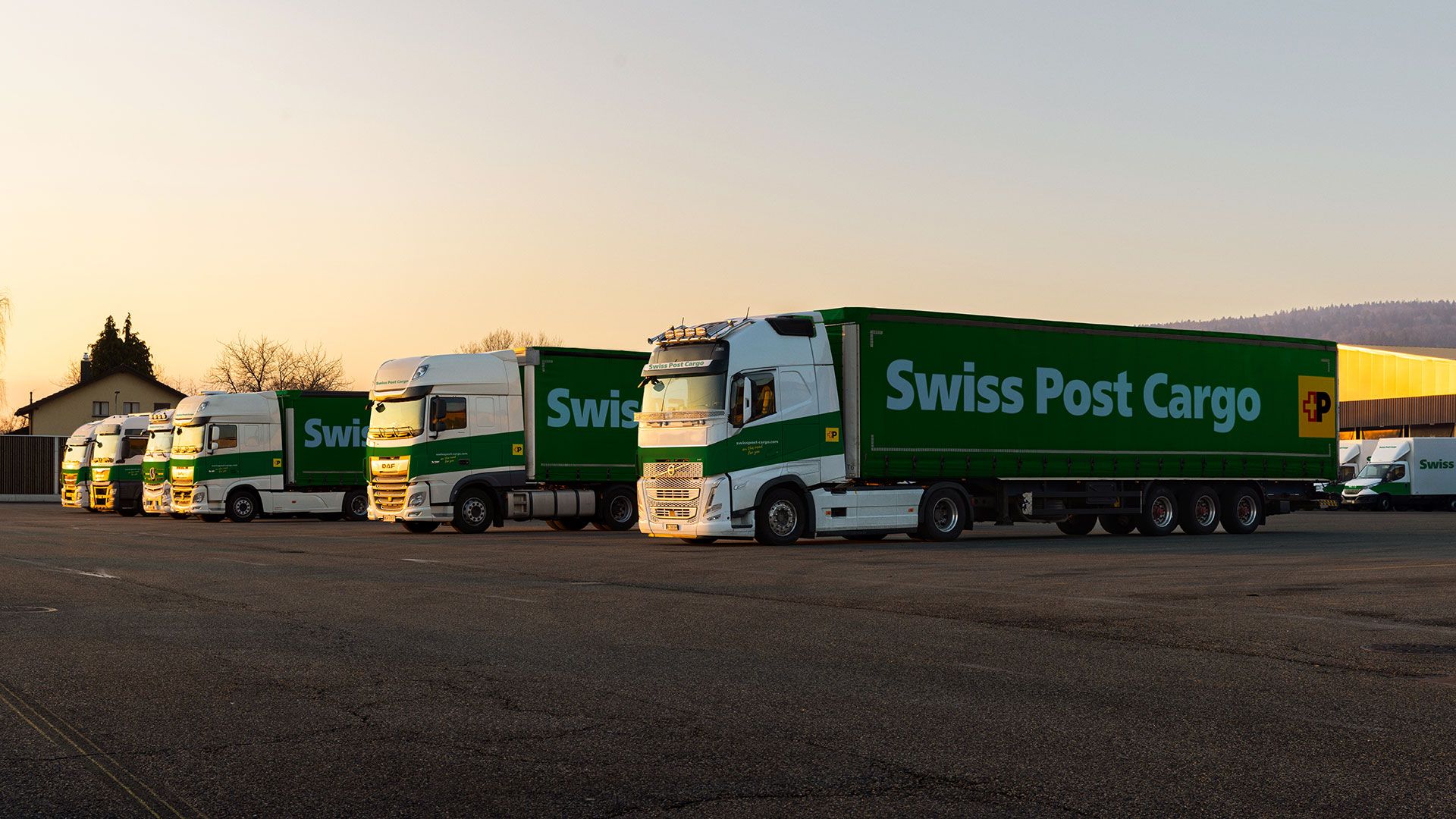 Swiss Post Cargo lorries stand in front of the logistics center at dusk.
