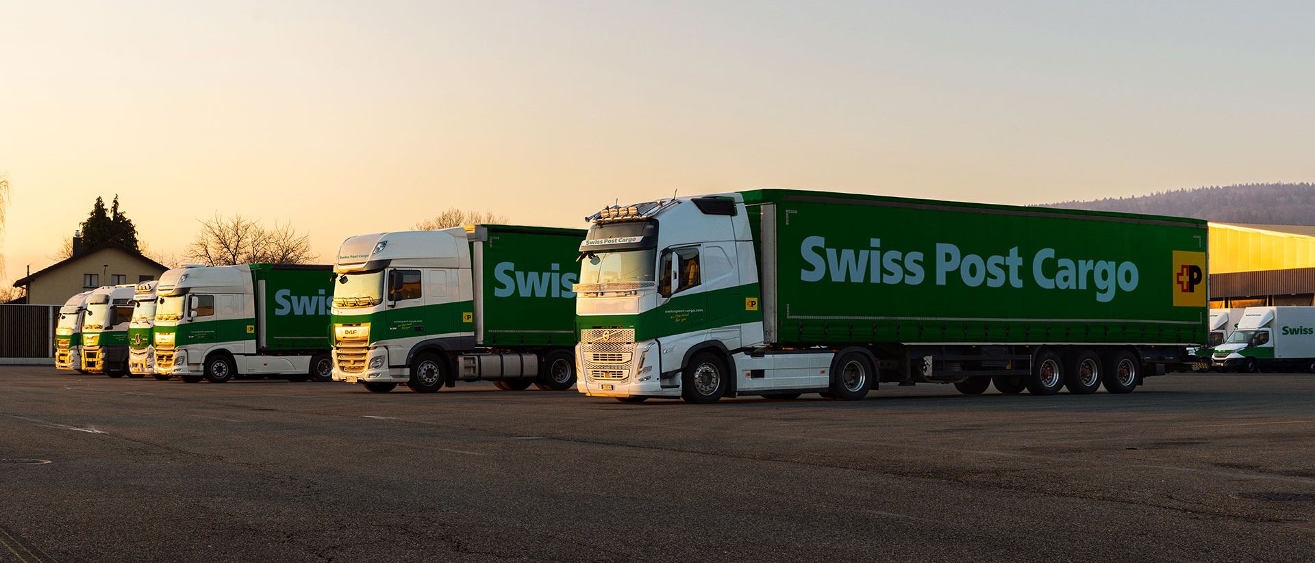 Swiss Post Cargo lorries stand in front of the logistics center at dusk.