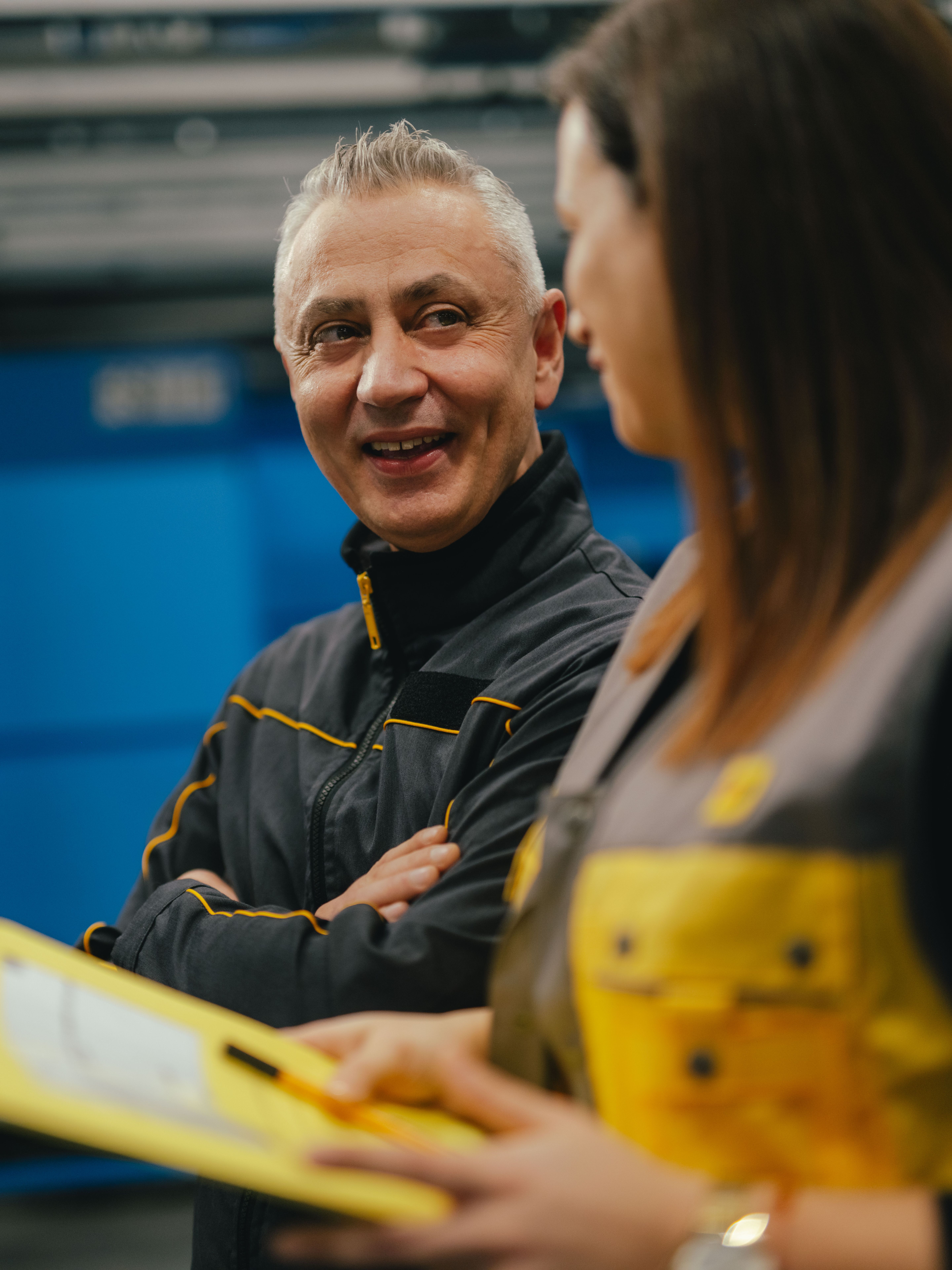 Two people are in an industrial setting, looking together at a yellow clipboard held by one of them. Both are wearing dark work clothing with yellow accents. In the background, blue machine housings, metal structures, and a fire safety station are visible.