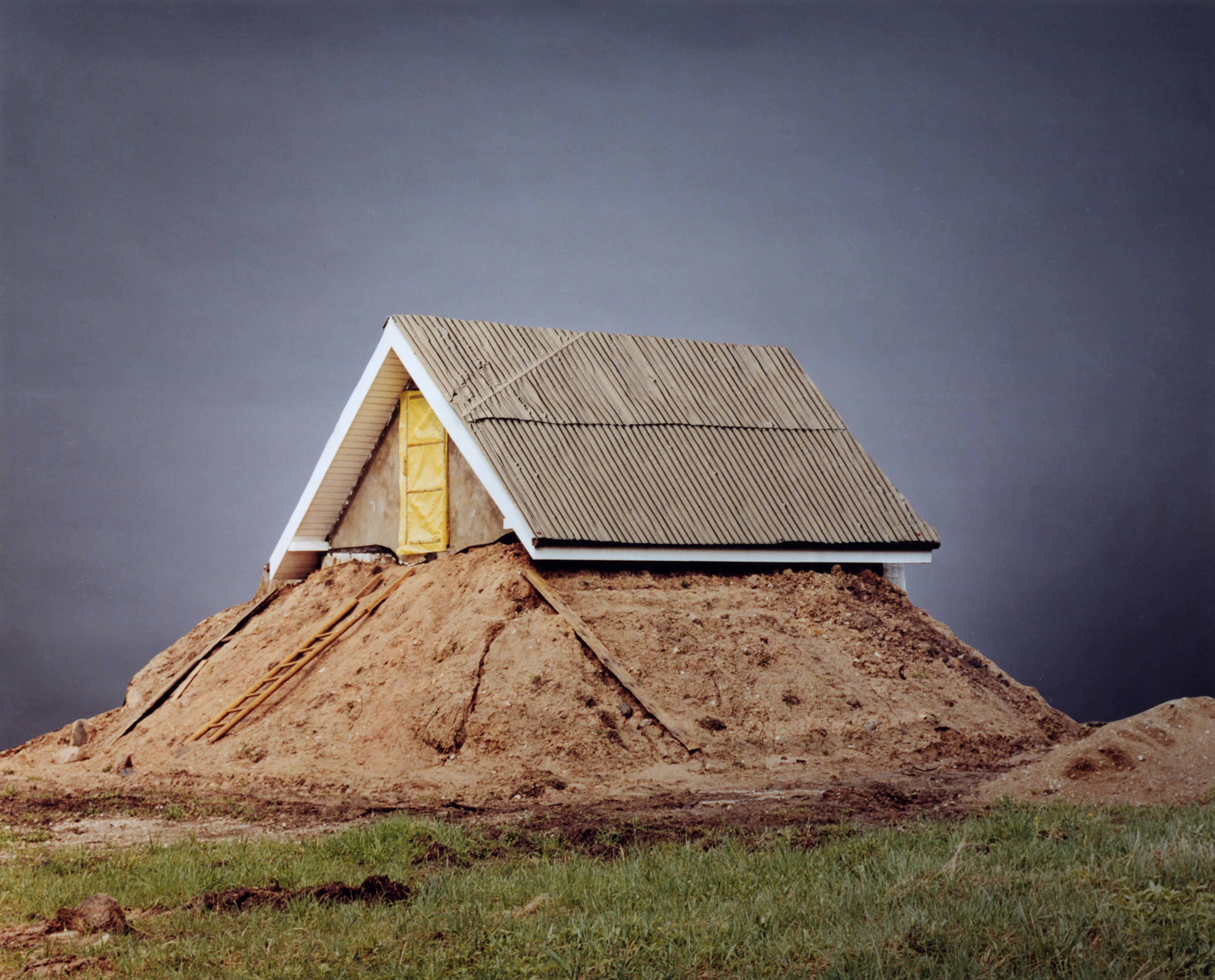 A small building with a corrugated iron roof stands on a mound against a grey background.