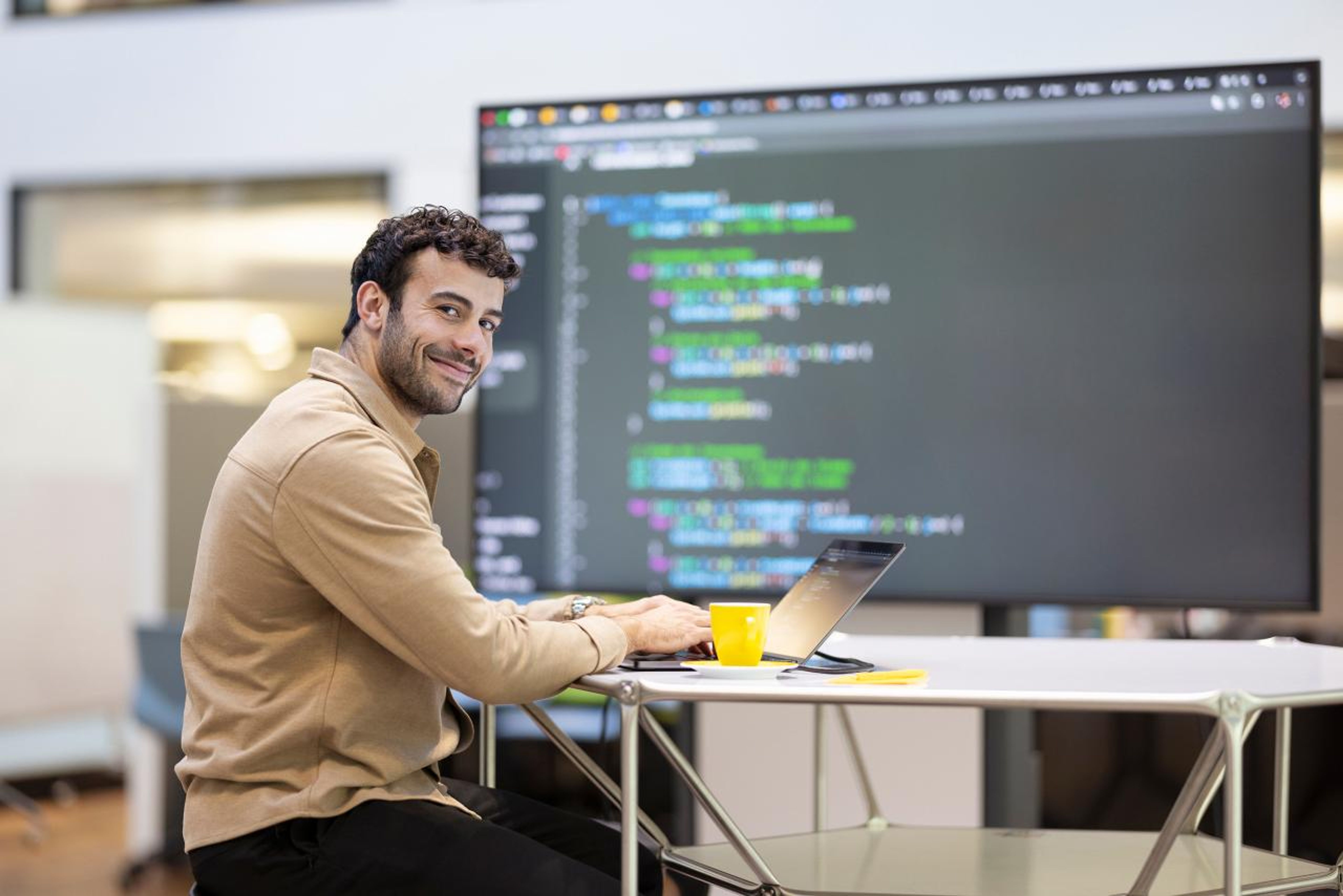 Ein Mann sitzt an einem Tisch und arbeitet an einem Laptop vor einem grossen Monitor, auf dem Code angezeigt wird. Er lächelt in die Kamera, die Umgebung wirkt modern und freundlich. Der Raum ist gut beleuchtet und die Atmosphäre wirkt professionell und kreativ.,