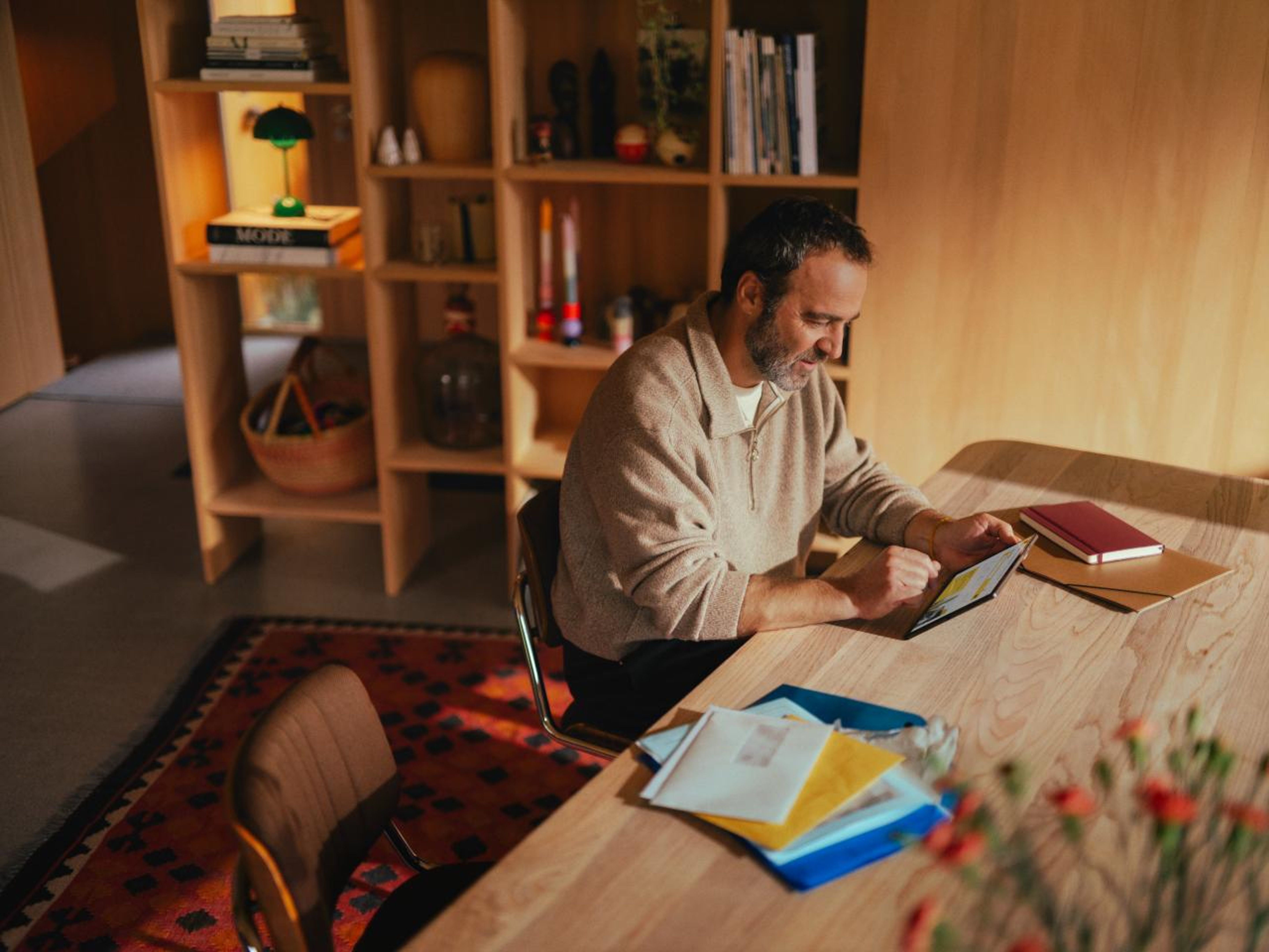 Ein Mann sitzt an einem Holztisch in einem gemütlichen Raum und verwendet ein Tablet. Auf dem Tisch liegen Briefe und ein Notizbuch. Der Hintergrund zeigt ein Regal mit Büchern und Dekorationen, was eine warme und einladende Atmosphäre schafft.