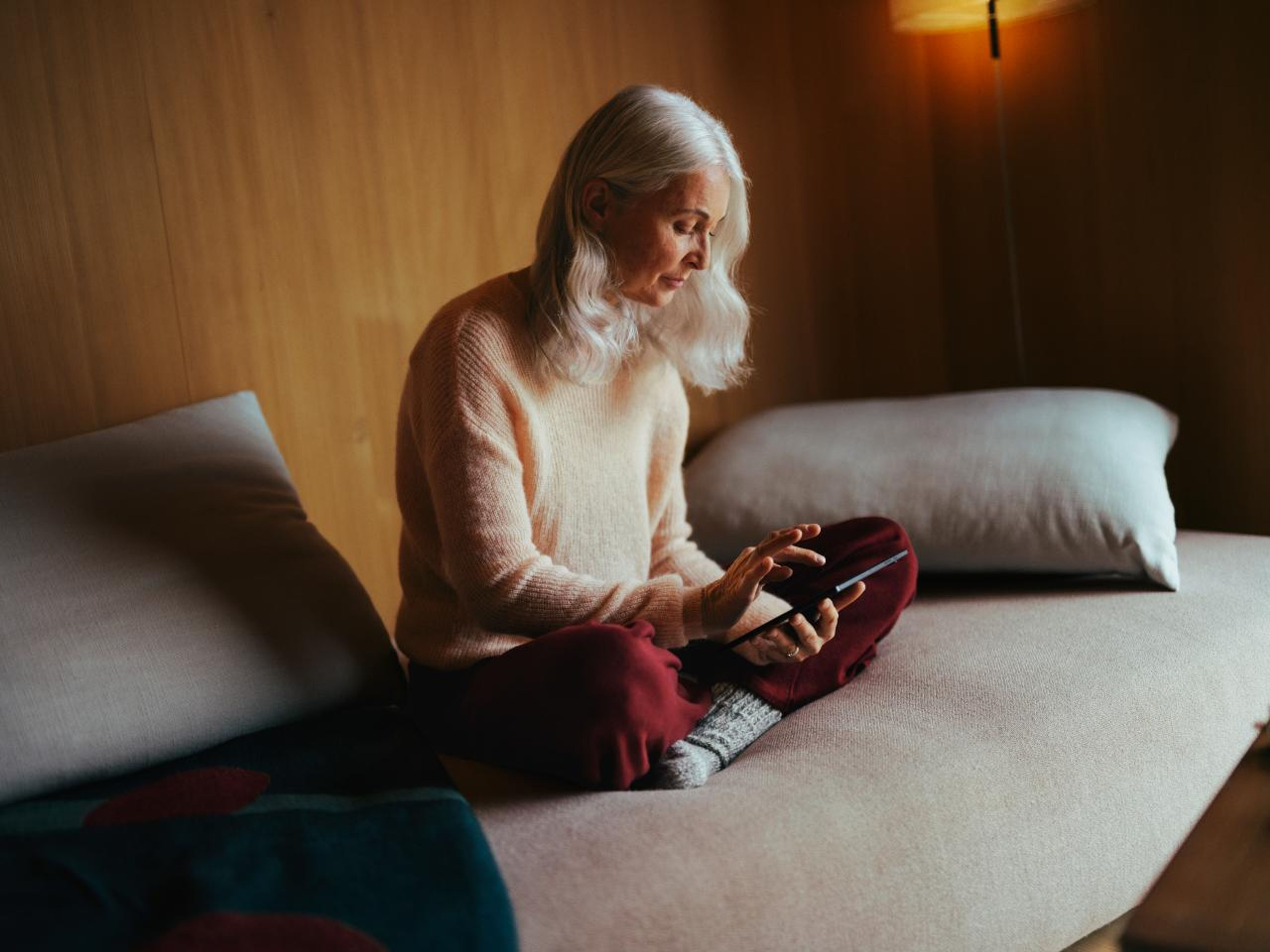 Eine ältere Frau sitzt bequem auf einem Sofa und bedient ein Tablet. Der Raum ist warm beleuchtet, mit einer Holzwand und sanfter Beleuchtung, was eine ruhige und entspannte Atmosphäre schafft.