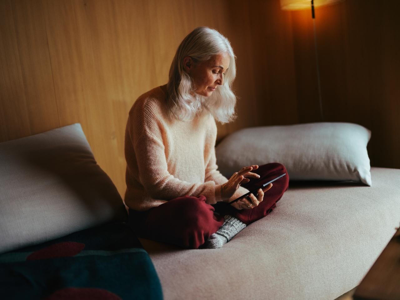 Eine ältere Frau sitzt bequem auf einem Sofa und bedient ein Tablet. Der Raum ist warm beleuchtet, mit einer Holzwand und sanfter Beleuchtung, was eine ruhige und entspannte Atmosphäre schafft.