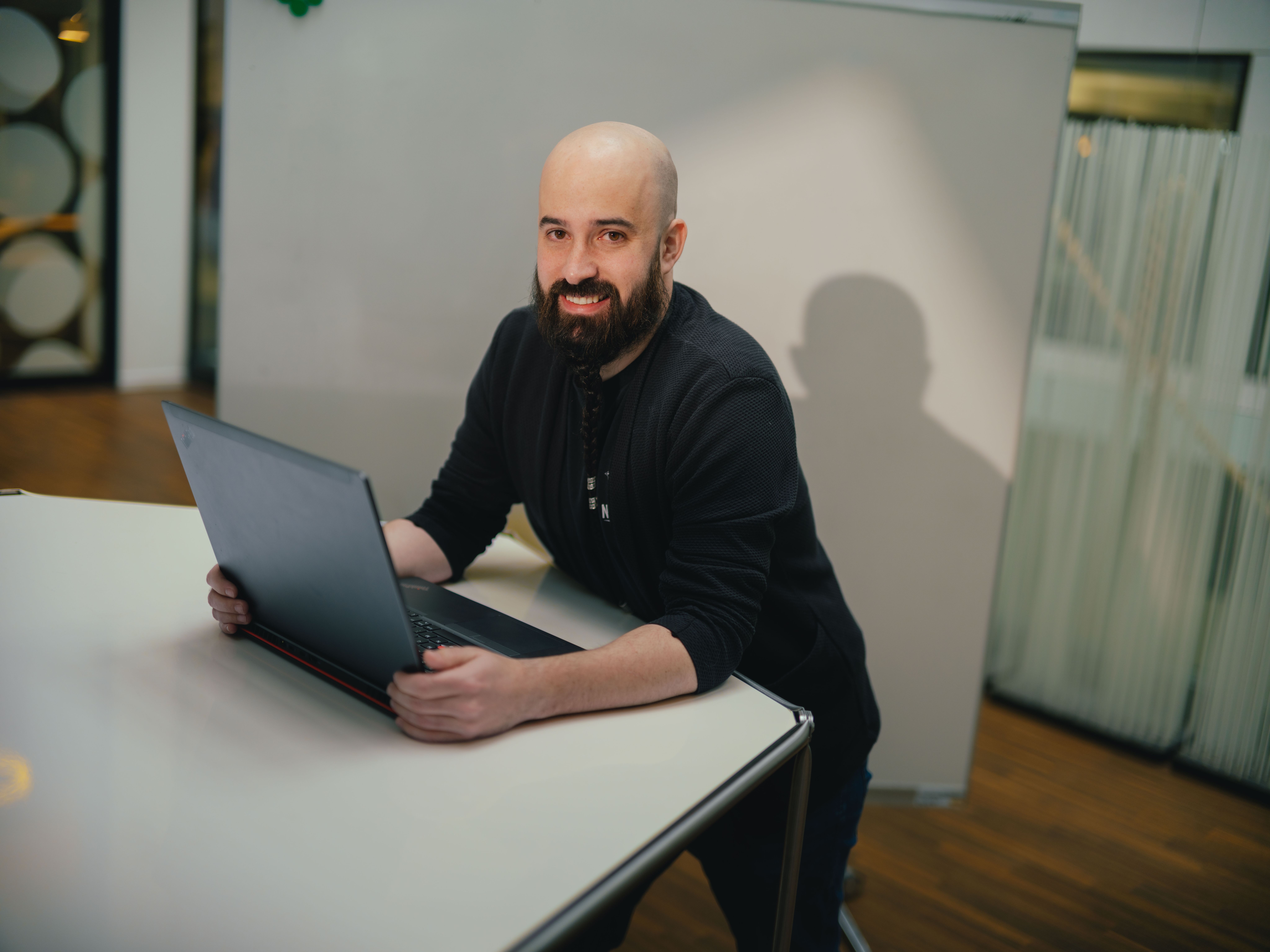 Eine Person sitzt an einem Tisch und arbeitet an einem aufgeklappten Laptop. Die Person trägt dunkle Kleidung. Im Hintergrund sind eine Glaswand mit vertikalen Strukturen sowie Teile eines weiteren Raumes erkennbar. Der Tisch ist hell und hat glatte Oberflächen.