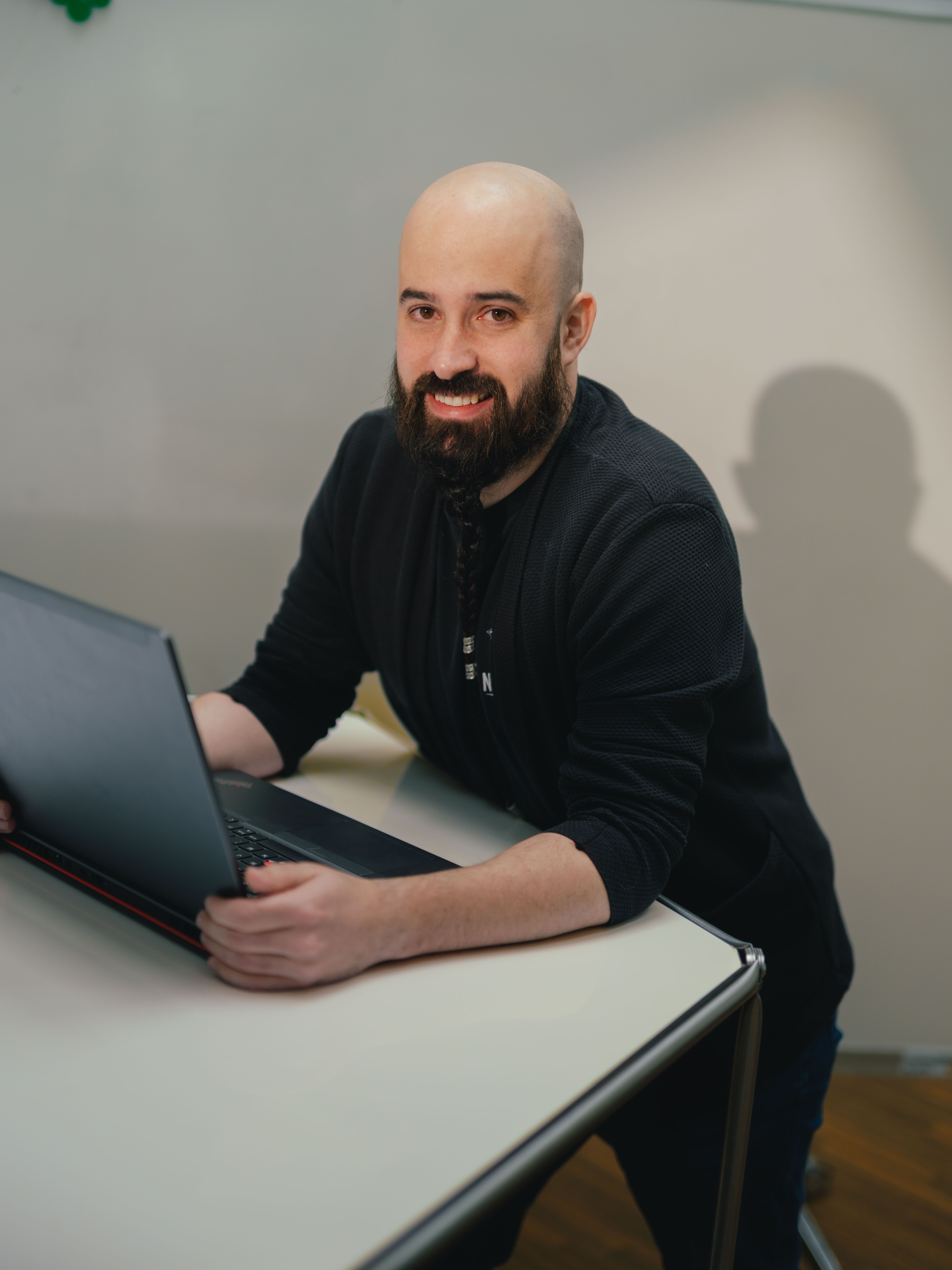 A person is seated at a table working on an open laptop. The person is wearing dark clothing. In the background, a glass wall with vertical structural elements and part of another room are visible. The table is light‑coloured with a smooth surface.