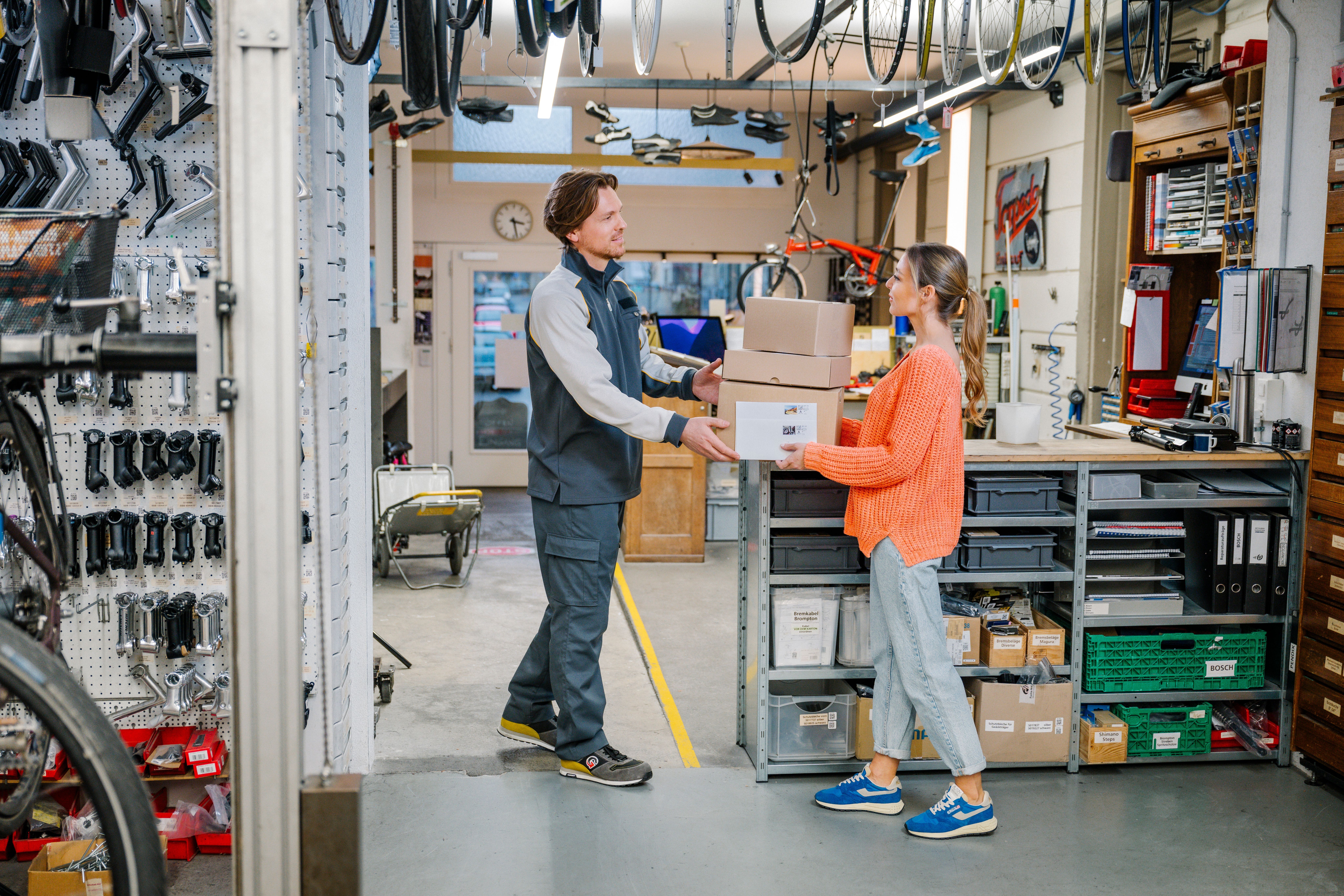One person hands over several parcels to another person in a workshop with bicycle accessories. Shelves with spare parts and tools are visible in the background.
