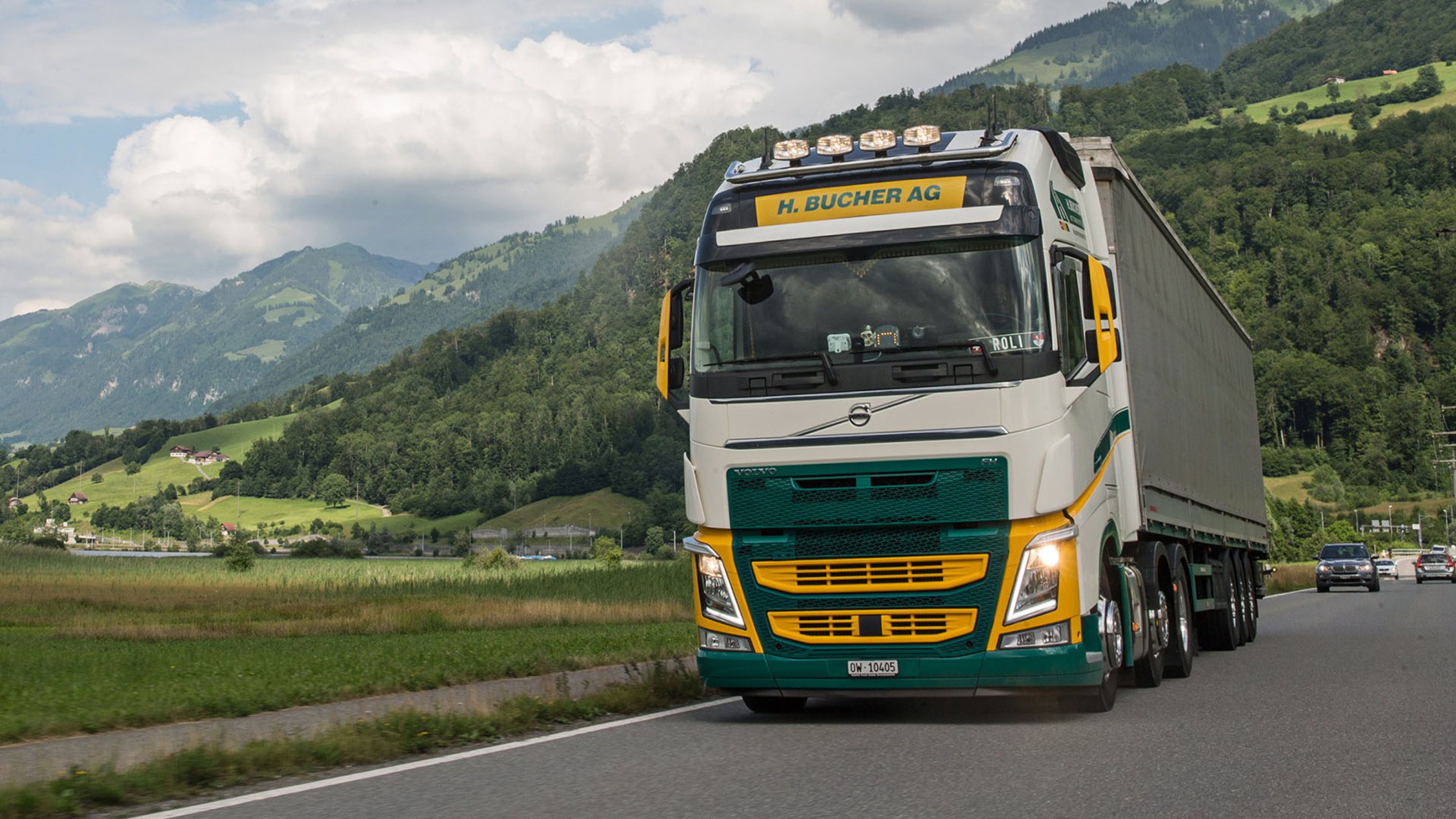 L’autocarro della H. Bucher AG percorre una strada di campagna attraverso un paesaggio alpino verdeggiante con le montagne sullo sfondo.
