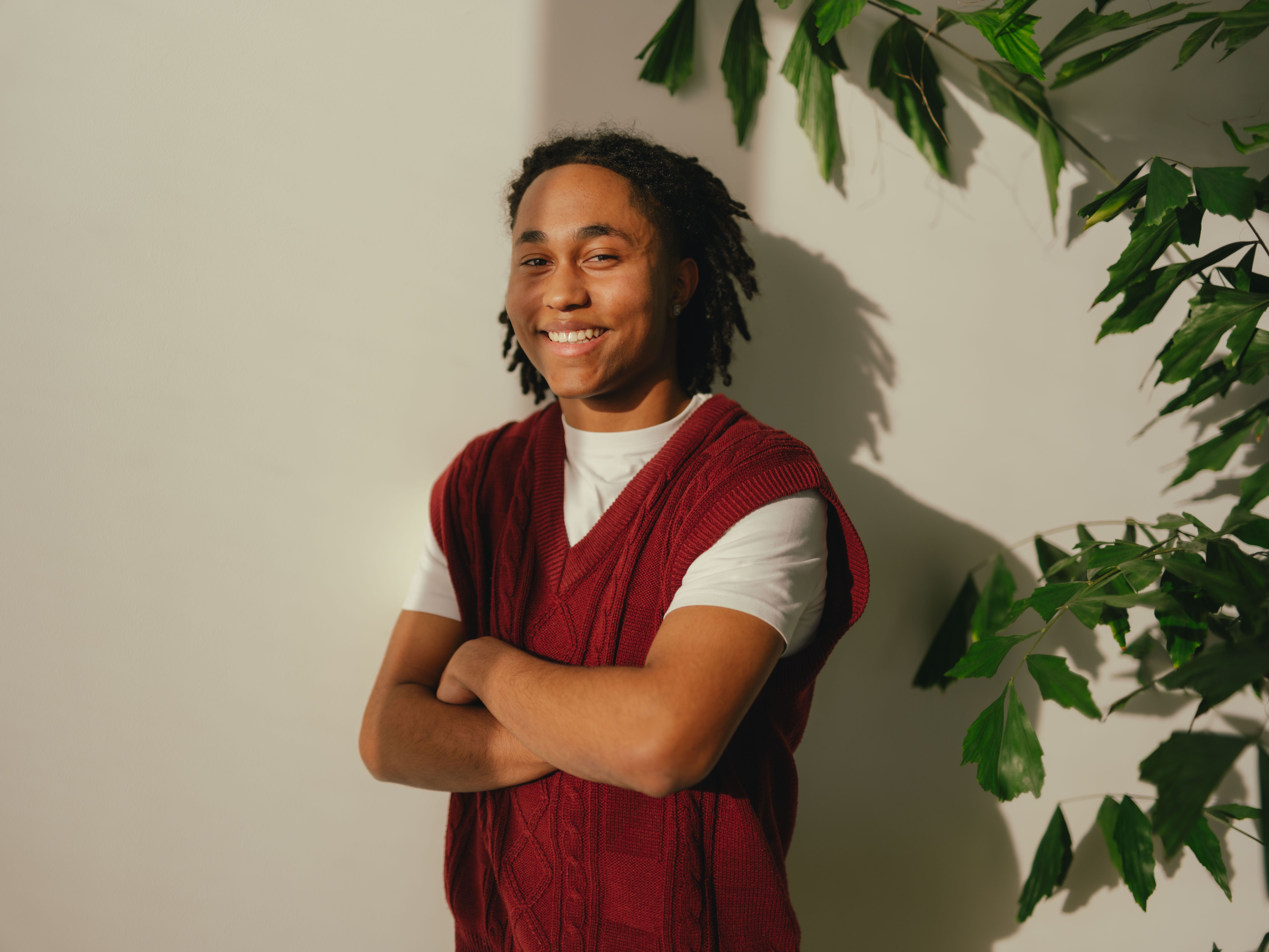 A person stands in front of a light‑coloured wall, partially framed by plant leaves and their shadows. The person is wearing a white T‑shirt under a red knitted sleeveless sweater and has their arms crossed.