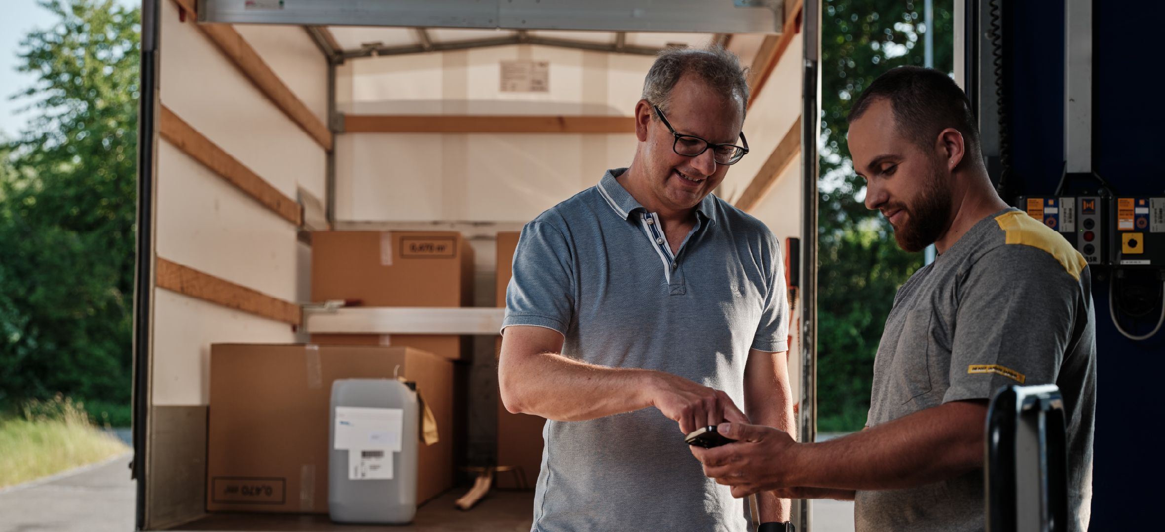 Two people stand by an open delivery van with several boxes inside. One points at a mobile device held by the other. Trees are visible in the background.