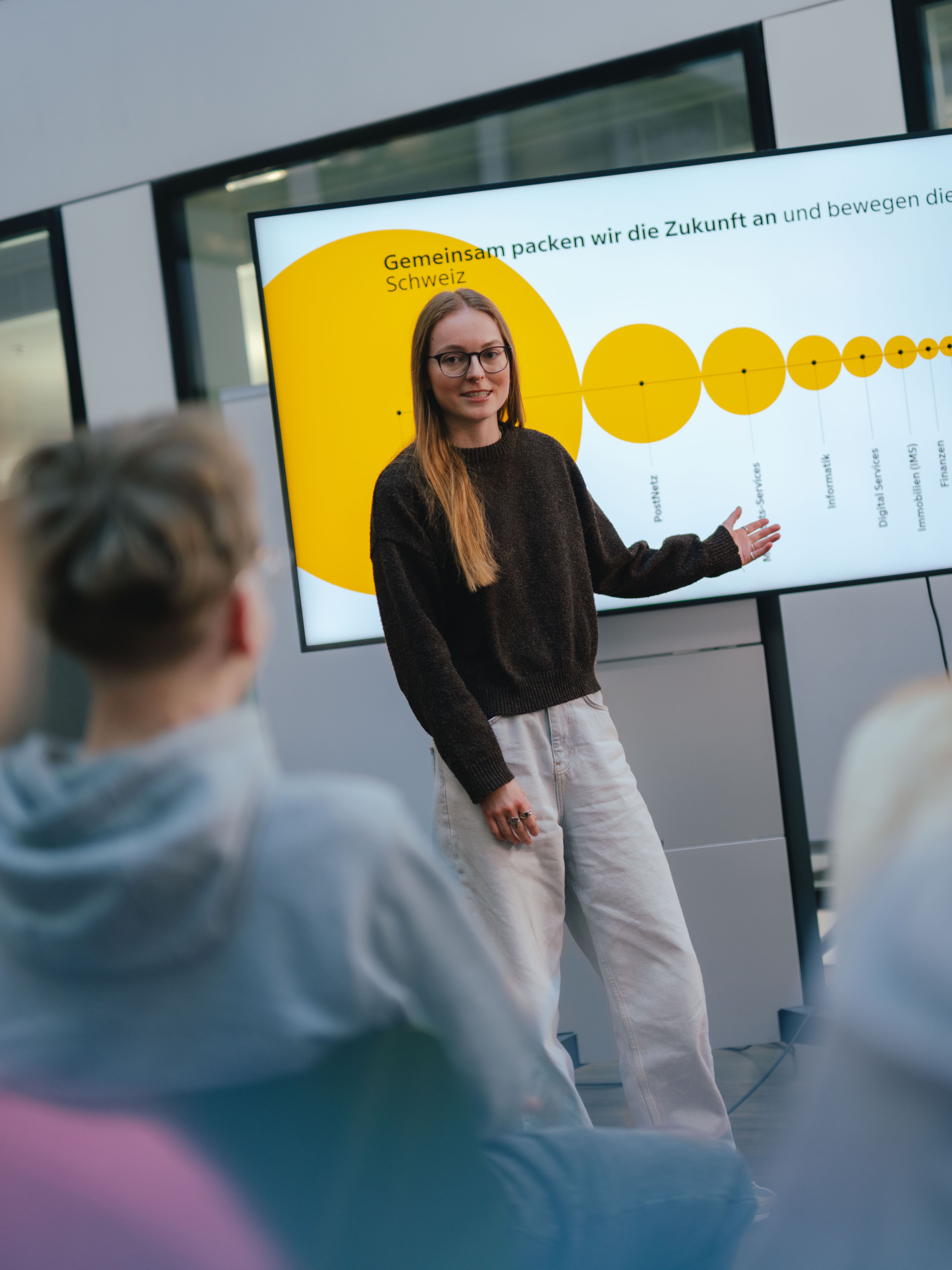Una persona è in piedi davanti a un grande schermo. Sullo schermo è visibile un grafico giallo con cerchi di diverse dimensioni, uniti in una linea. Diverse altre persone siedono davanti alla persona in piedi e guardano la presentazione. L’aula ha l’aspetto di una moderna sala riunioni o un’aula di formazione.