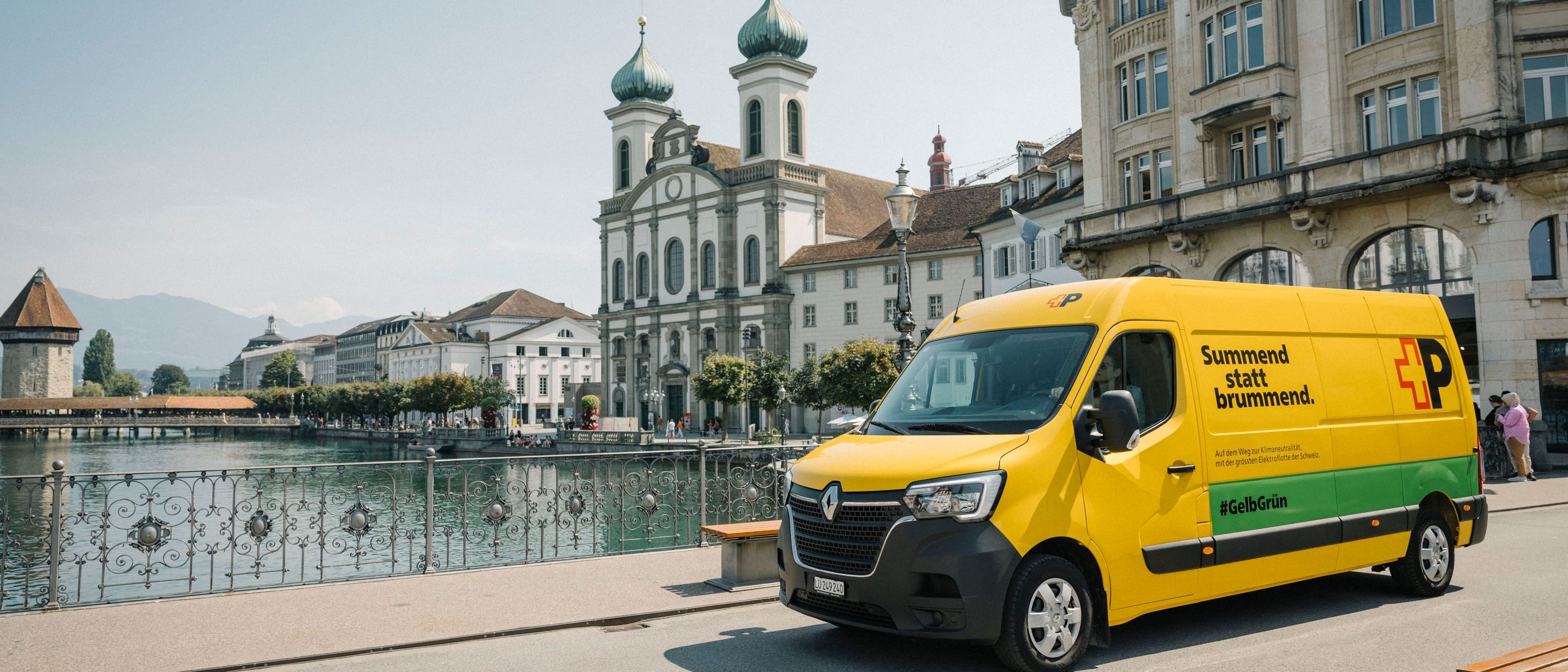 A yellow Swiss Post delivery van is parked on a riverside promenade in Lucerne. In the background are the Jesuit Church, historic buildings, and the Reuss River. Text on the van reads: “Summend statt brummend.”.