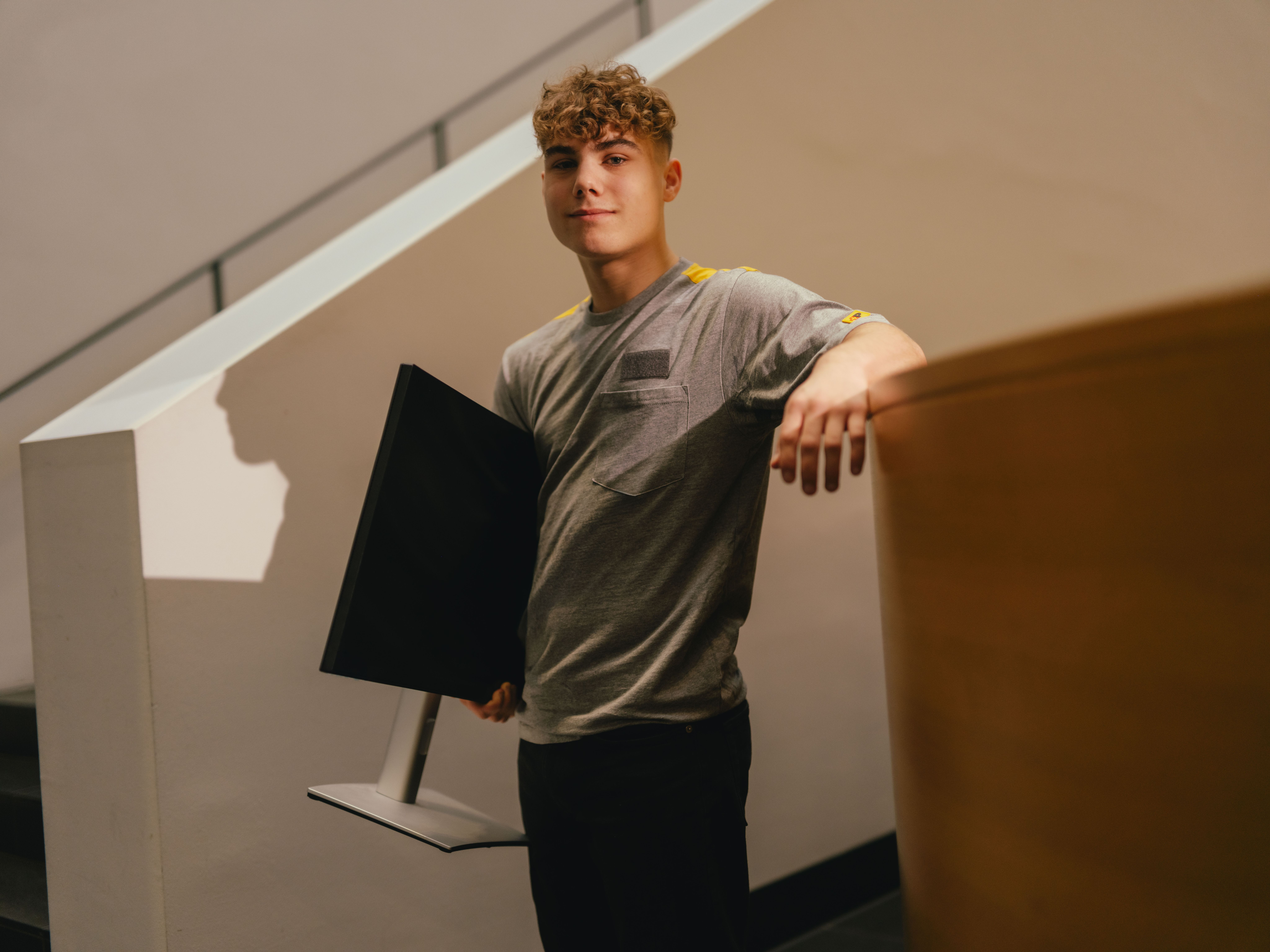 Une personne se tient en intérieur à côté d’un escalier clair et s’appuie avec un bras sur une surface surélevée. La personne porte un t-shirt gris et tient sous l’autre bras un écran de couleur foncée. L’arrière-plan montre des lignes architecturales claires et une lumière douce et chaude.
