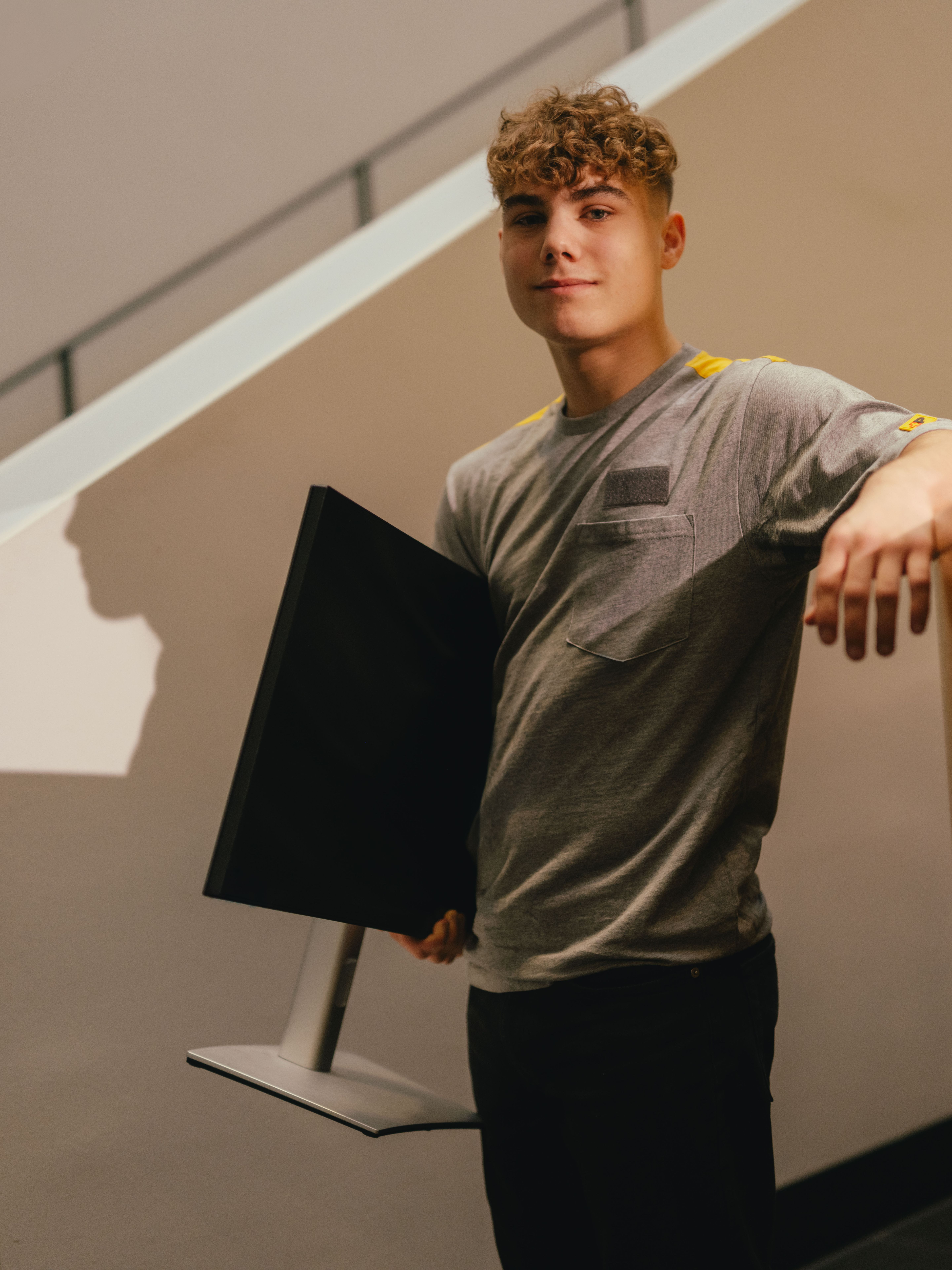 Une personne se tient en intérieur à côté d’un escalier clair et s’appuie avec un bras sur une surface surélevée. La personne porte un t-shirt gris et tient sous l’autre bras un écran de couleur foncée. L’arrière-plan montre des lignes architecturales claires et une lumière douce et chaude.