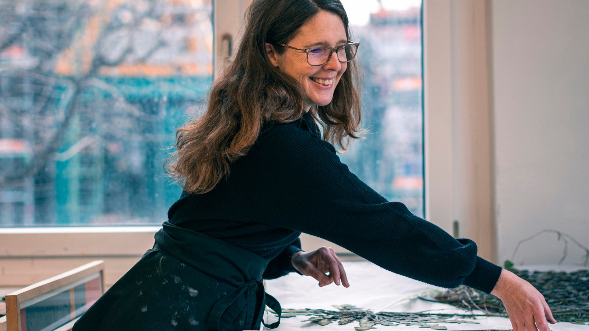 Eine Frau mit Brille in schwarzem Pullover und Schürze arbeitet an einem Tisch vor einem Fenster.