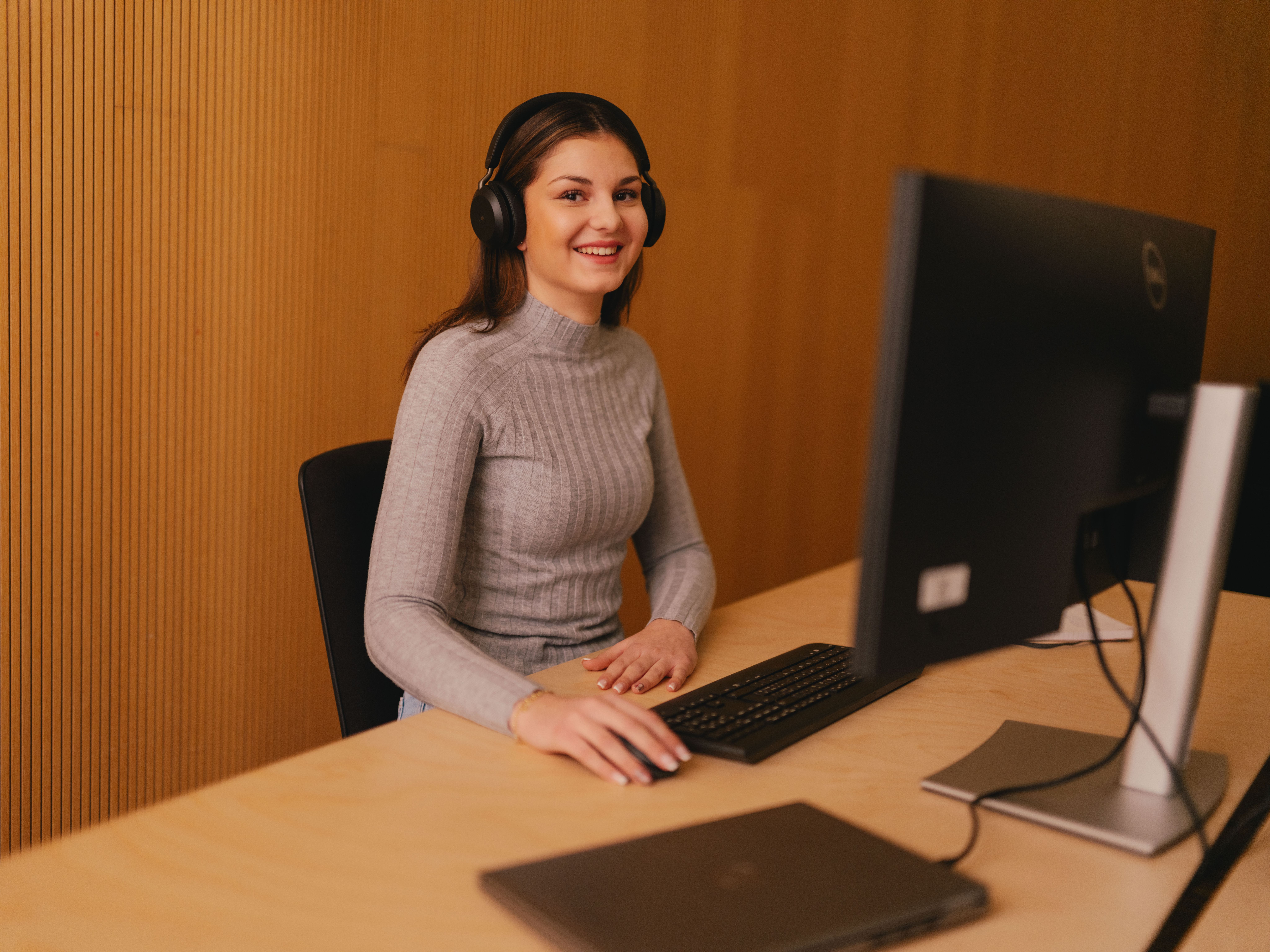 Eine Person sitzt an einem Schreibtisch und arbeitet an einem Desktop‑Computer. Die Person trägt grosse Over‑Ear‑Kopfhörer und ein geripptes, langärmeliges Oberteil. Vor der Person stehen eine Tastatur und eine Computermaus. Auf dem Schreibtisch liegt zudem ein geschlossenes Laptopgerät. Der Hintergrund besteht aus senkrechten Holzpaneelen.