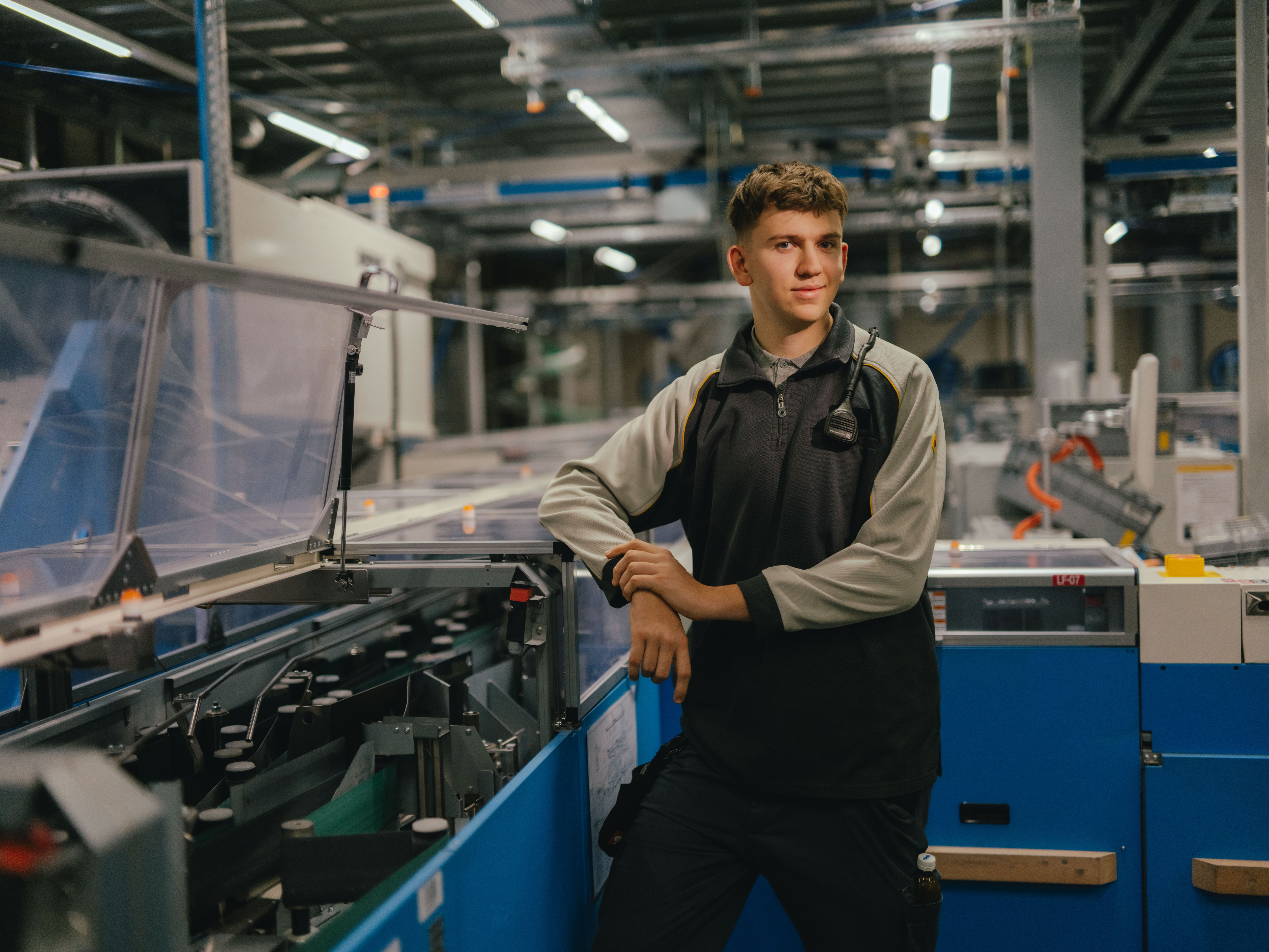 Eine Person steht in einer industriellen Halle und lehnt sich mit beiden Armen auf eine Maschine mit transparenten Abdeckungen. Die Person trägt eine zweifarbige Arbeitsjacke. Im Hintergrund sind weitere Maschinen, Metallstrukturen und Beleuchtungselemente der Produktionsumgebung sichtbar.