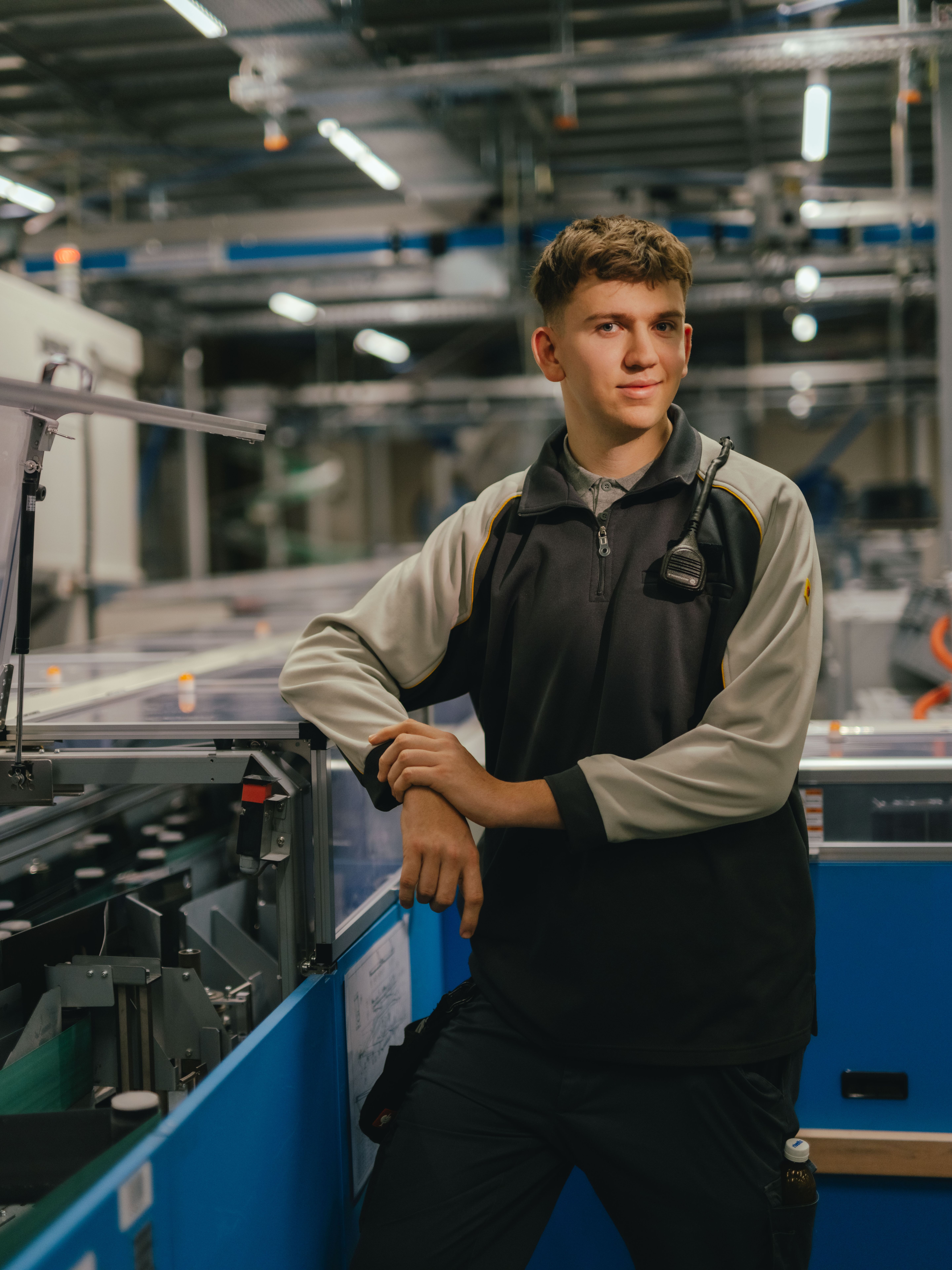 A person is standing in an industrial hall and leaning with both arms on a machine with transparent covers. They are wearing a two-coloured work jacket. Other machines, metal structures and lighting elements from the production environment can be seen in the background.