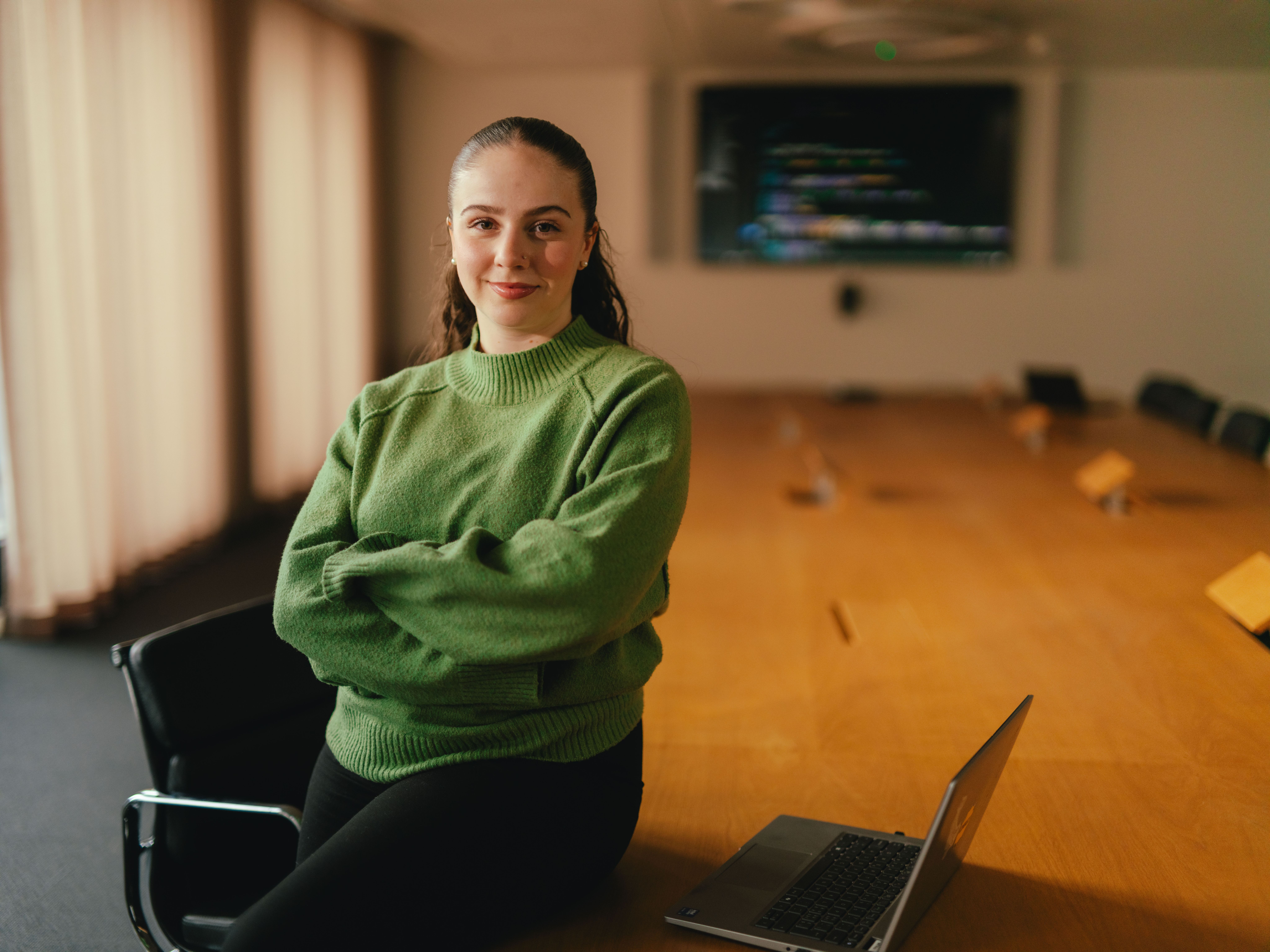 A person is standing in a conference room next to a large wooden table, arms loosely folded in front of their body. The person is wearing a green pullover. In the background, there is a large screen on the wall, several chairs and large windows with closed curtains. There is an open laptop on the table.
