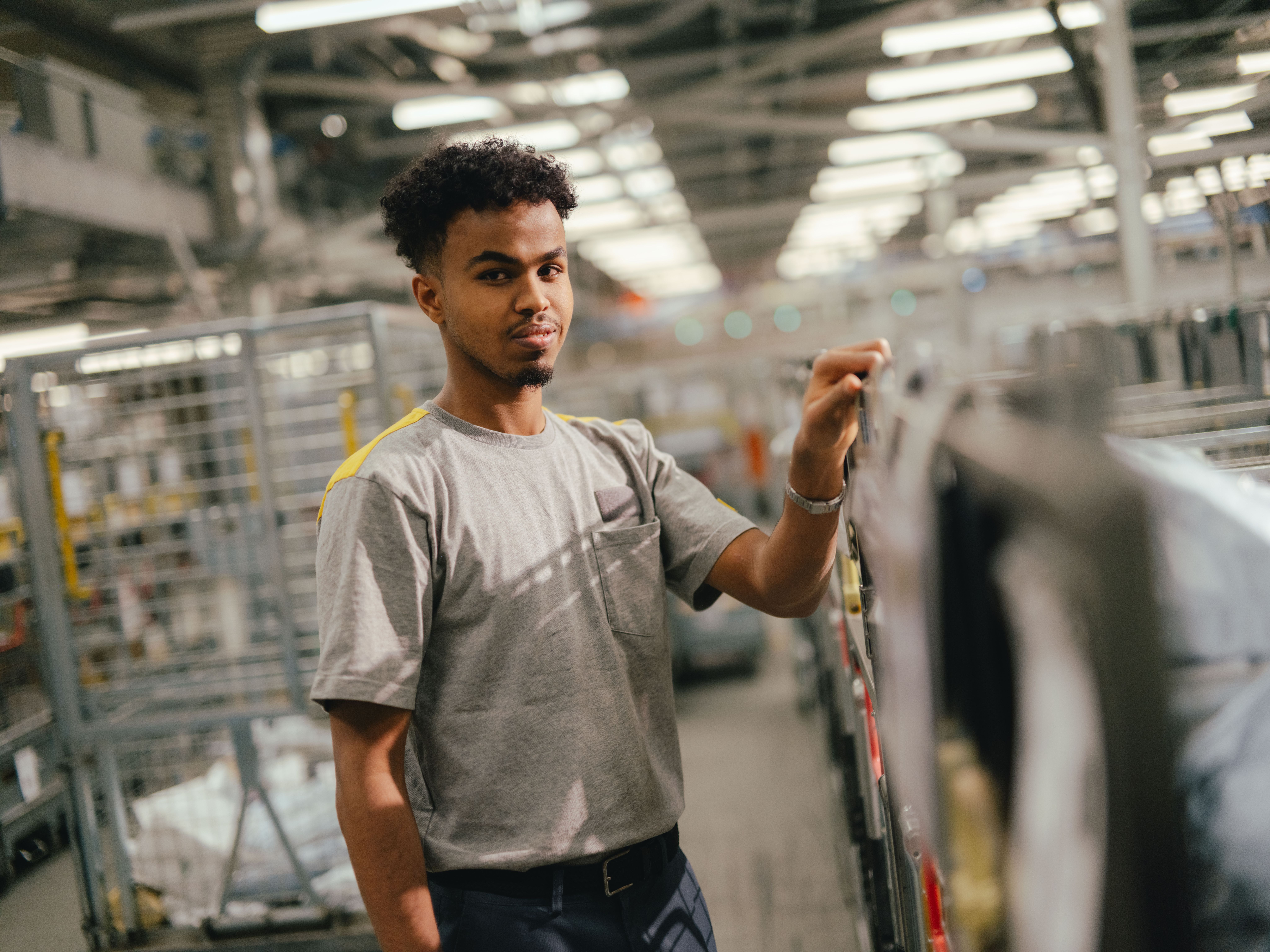 Eine Person steht in einer grossen Halle zwischen Metallregalen und Transportwagen. Die Person trägt ein helles T‑Shirt und hält mit einer Hand einen Handgriff an einem Wagen oder Gestell. Die Halle ist hell beleuchtet und zeigt zahlreiche industrielle Strukturen im Hintergrund.