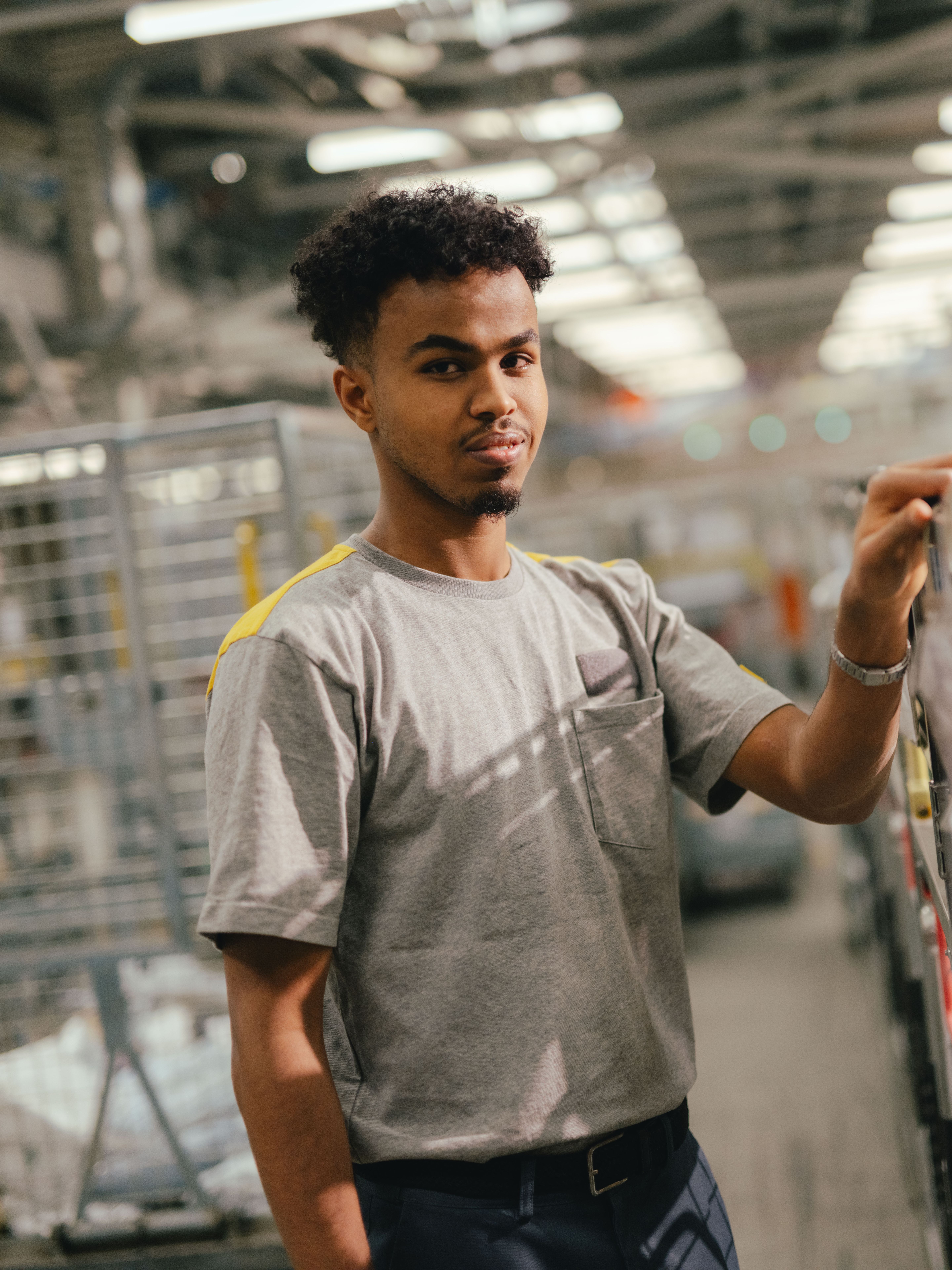 Eine Person steht in einer grossen Halle zwischen Metallregalen und Transportwagen. Die Person trägt ein helles T‑Shirt und hält mit einer Hand einen Handgriff an einem Wagen oder Gestell. Die Halle ist hell beleuchtet und zeigt zahlreiche industrielle Strukturen im Hintergrund.