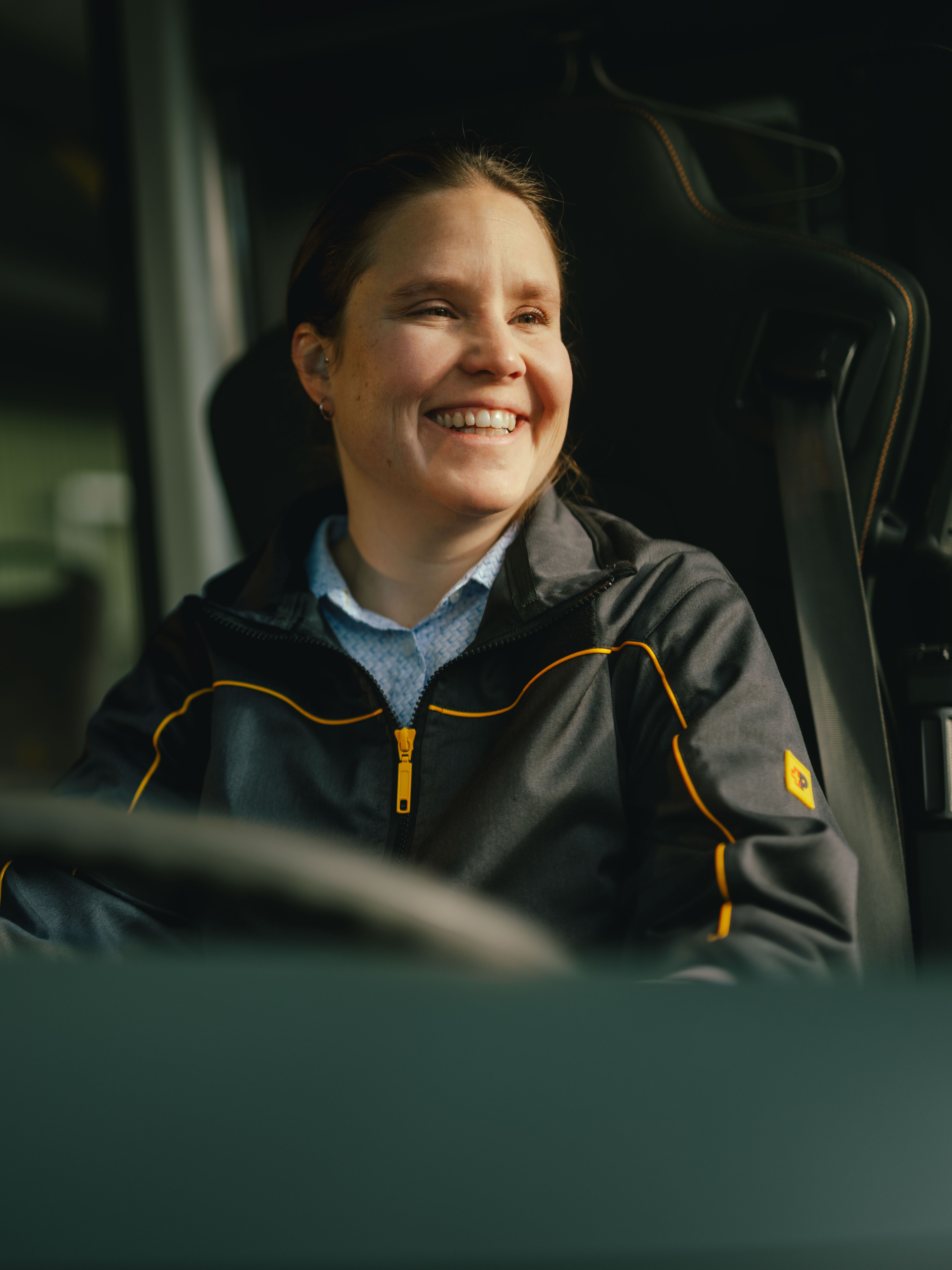 Une personne est assise au volant d’un véhicule, portant une veste foncée avec des accents jaunes. L’intérieur du véhicule, y compris le volant et des poignées, est visible.