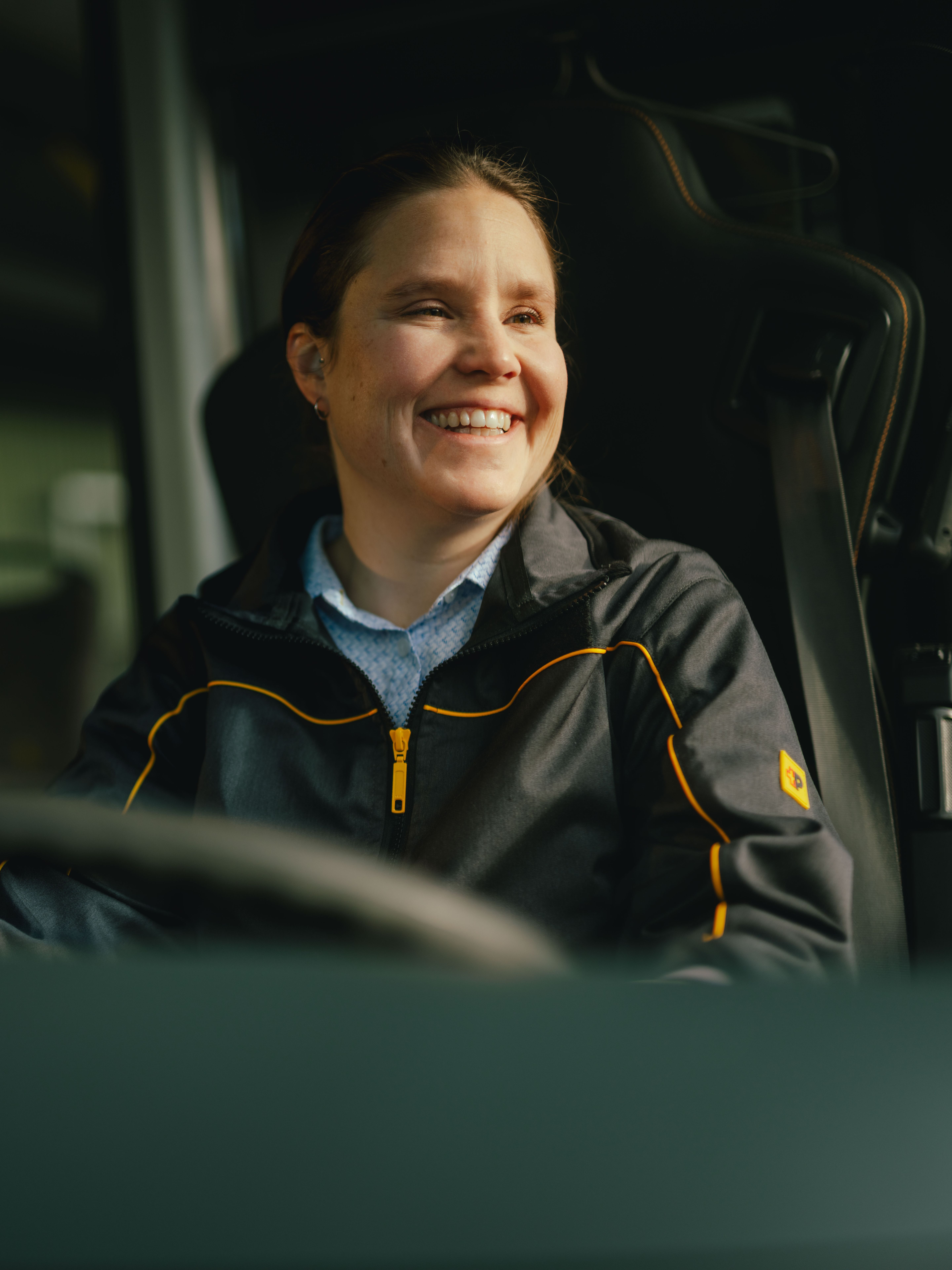 Eine Person sitzt am Steuer eines Fahrzeugs und trägt eine dunkle Jacke mit gelben Akzenten. Teile des Innenraums des Fahrzeugs, einschliesslich des Lenkrads und Haltegriffen, sind sichtbar.