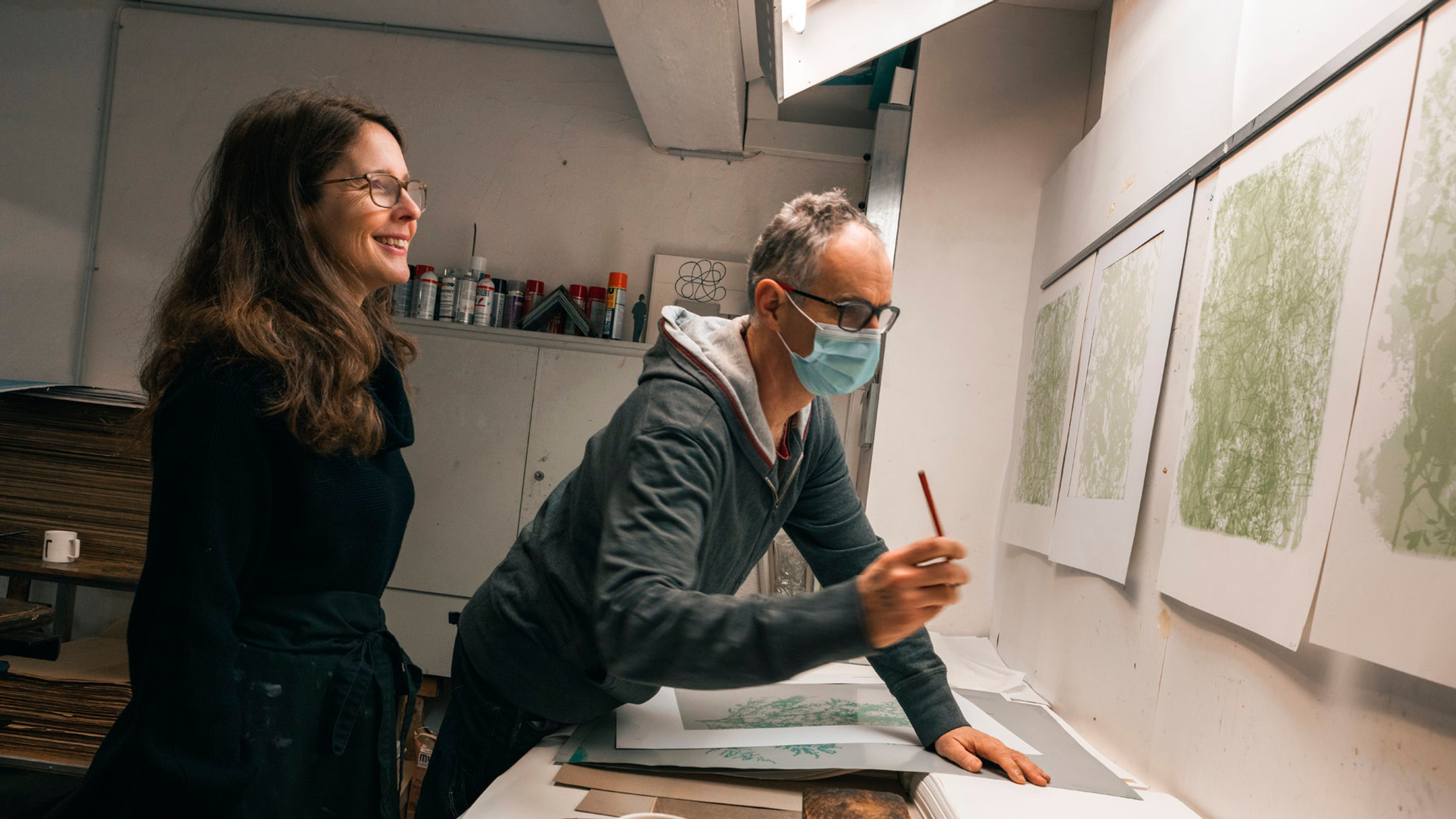 Eine Frau und ein Mann arbeiten in einem Atelier an Kunstwerken, während vier Bilder an der Wand hängen.
