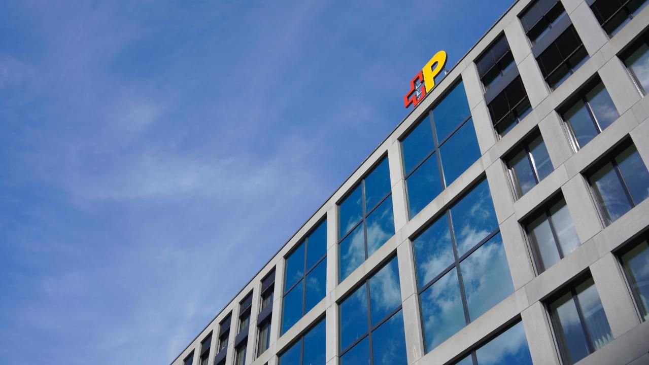 Das Bild zeigt die Fassade eines modernen Bürogebäudes mit grossen Glasfenstern und einem Logo der Schweizerischen Post auf dem Dach. Der Himmel ist klar und blau, was die Architektur des Gebäudes unterstreicht.,