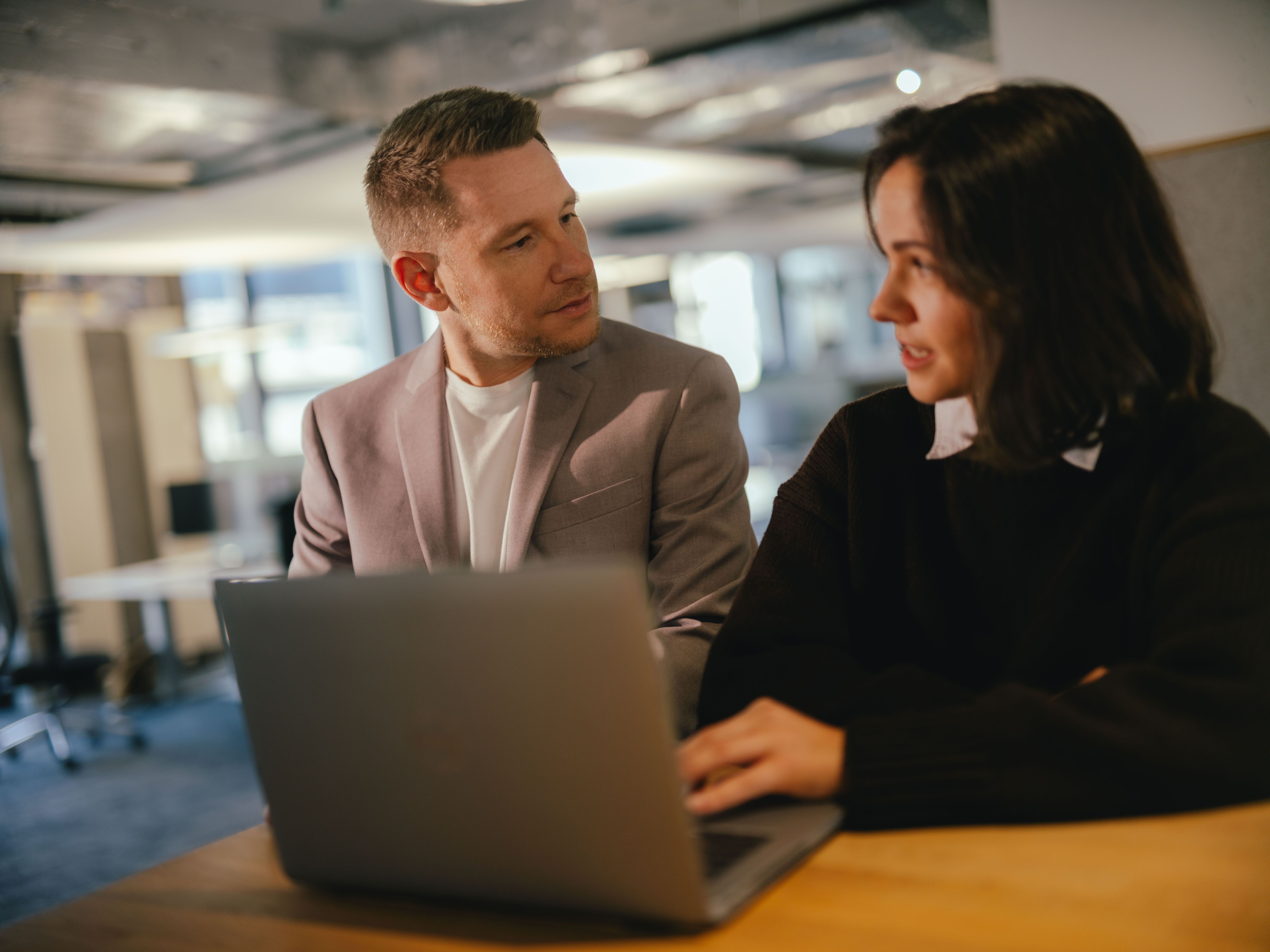 Zwei Personen sitzen an einem Tisch in einem modernen Büro und arbeiten gemeinsam an einem Laptop. Eine der Personen tippt auf der Tastatur, während die andere Person daneben sitzt und in Richtung des Bildschirms schaut. Im Hintergrund sind grosse Fenster und Büroeinrichtungen sichtbar.