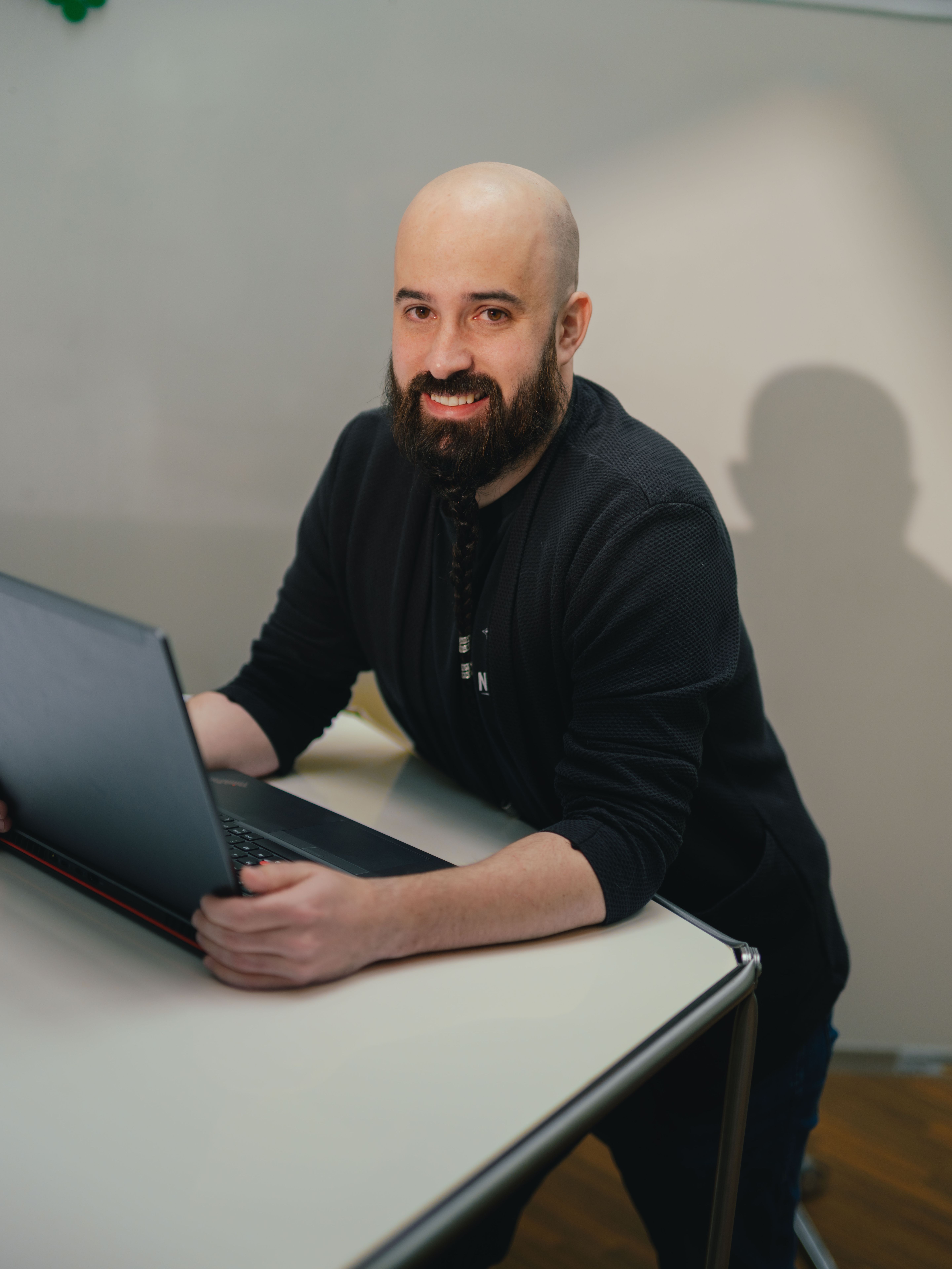 Una persona è seduta a un tavolo e lavora su un computer portatile aperto. La persona indossa abiti scuri. Sullo sfondo si vede una parete di vetro con elementi verticali e una parte di un altro ambiente. Il tavolo è chiaro e ha una superficie liscia.