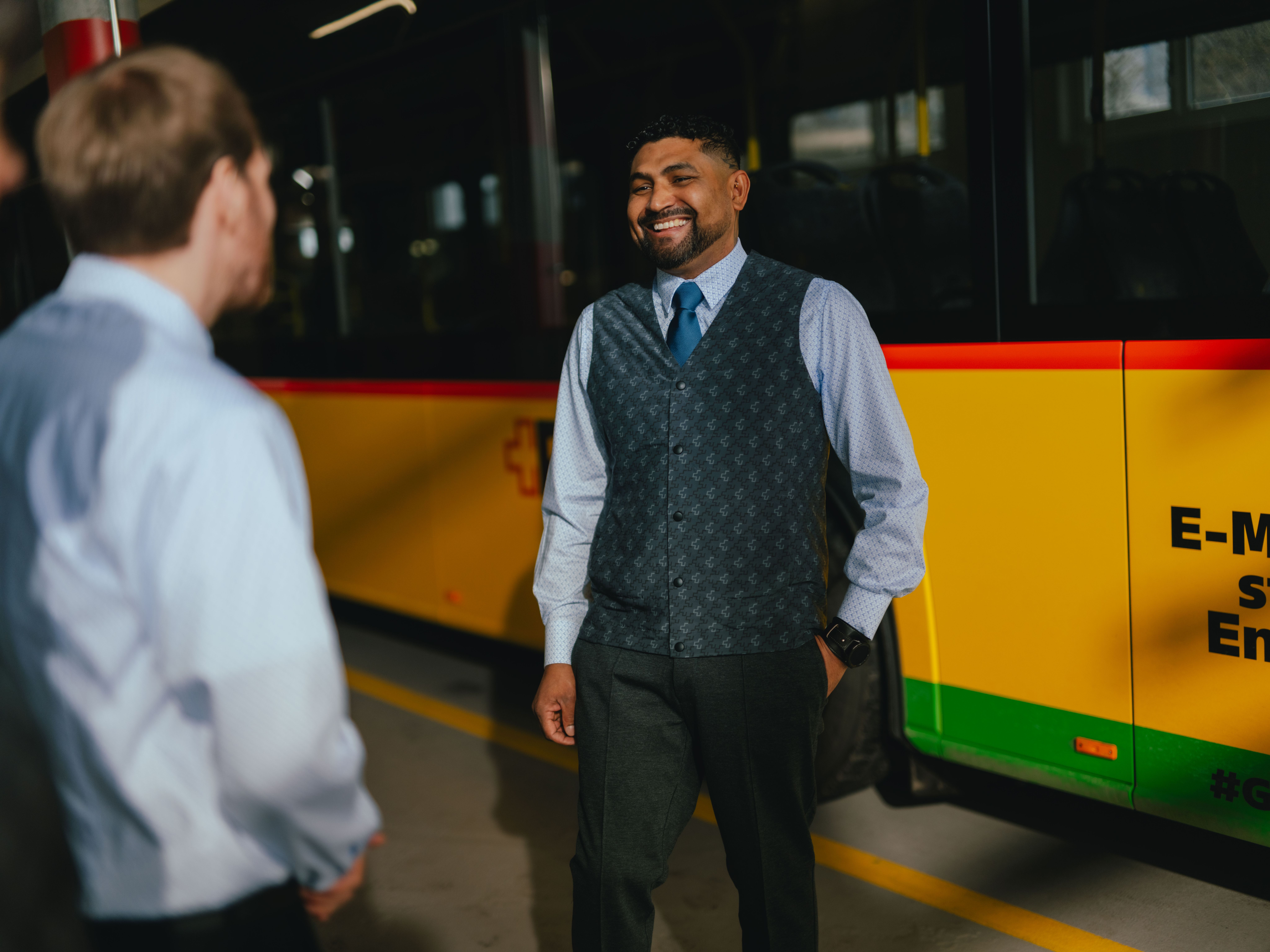 Une personne se tient dans un hall à côté d’un bus jaune et noir portant un texte publicitaire sur le côté. Une autre personne se tient en face et semble engagée dans une conversation. Les deux personnes portent des vêtements formels. Le sol comporte des marquages jaunes et l’environnement évoque un dépôt ou un espace de maintenance.