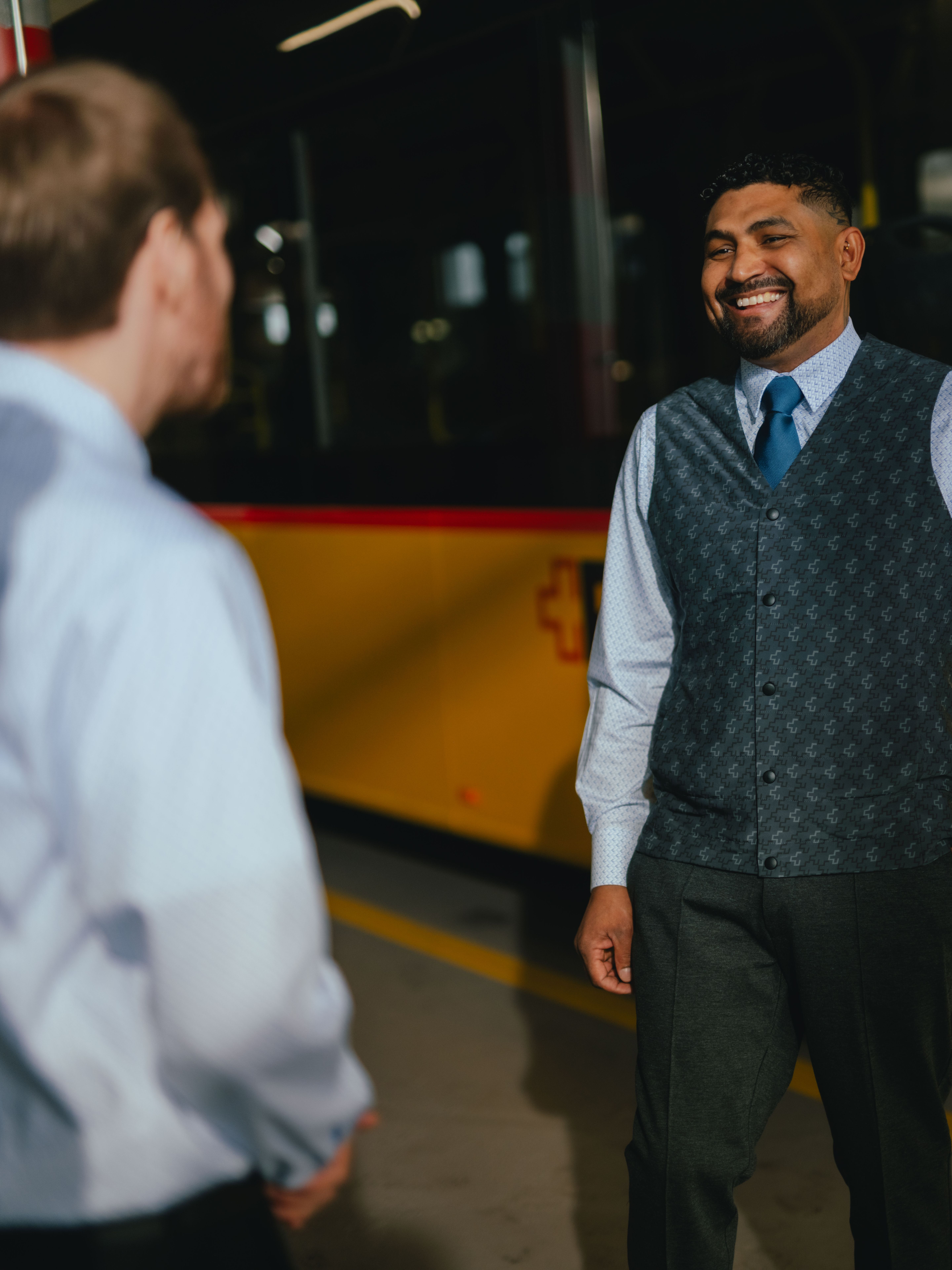 Une personne se tient dans un hall à côté d’un bus jaune et noir portant un texte publicitaire sur le côté. Une autre personne se tient en face et semble engagée dans une conversation. Les deux personnes portent des vêtements formels. Le sol comporte des marquages jaunes et l’environnement évoque un dépôt ou un espace de maintenance.
