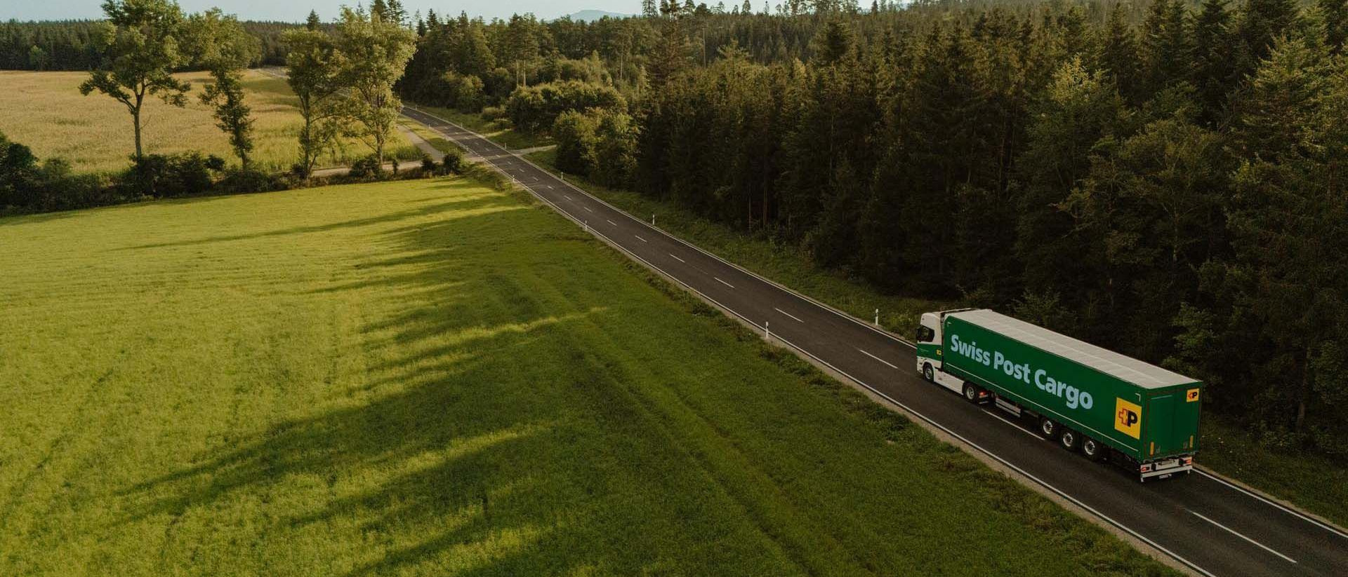 Un camion Swiss Post Cargo circule sur une route de campagne entre une prairie et une forêt.