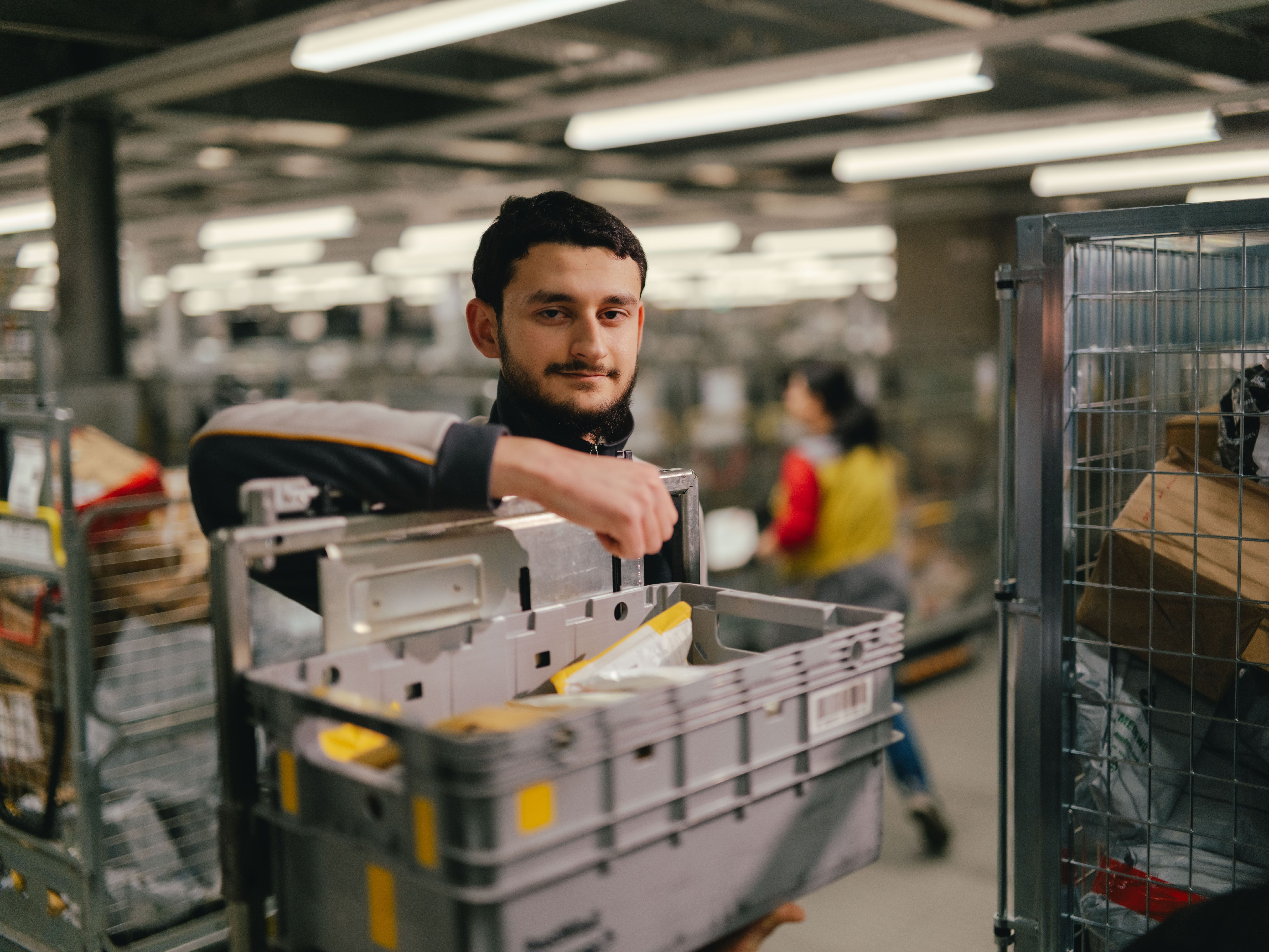 Eine Person steht in einer grossen Halle und hebt eine graue Kunststoffbox aus einem Metallrollwagen. In der Box befinden sich verschiedene Sendungen und Papiere. Der Arbeitsbereich ist mit vielen weiteren Wagen, Regalen und Beleuchtungselementen strukturiert, und im Hintergrund sind weitere Personen und Sortierstationen erkennbar.