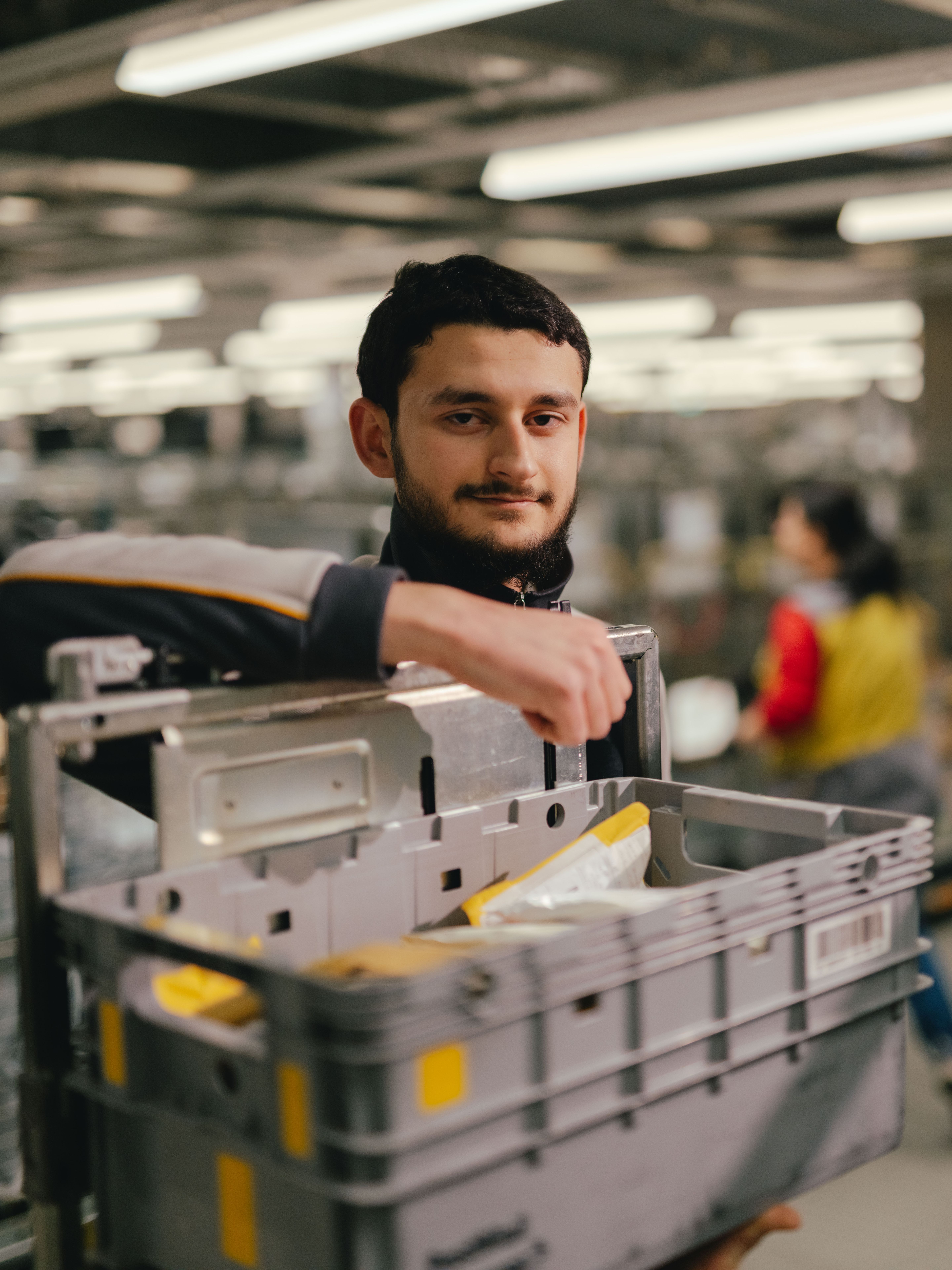 Eine Person steht in einer grossen Halle und hebt eine graue Kunststoffbox aus einem Metallrollwagen. In der Box befinden sich verschiedene Sendungen und Papiere. Der Arbeitsbereich ist mit vielen weiteren Wagen, Regalen und Beleuchtungselementen strukturiert, und im Hintergrund sind weitere Personen und Sortierstationen erkennbar.