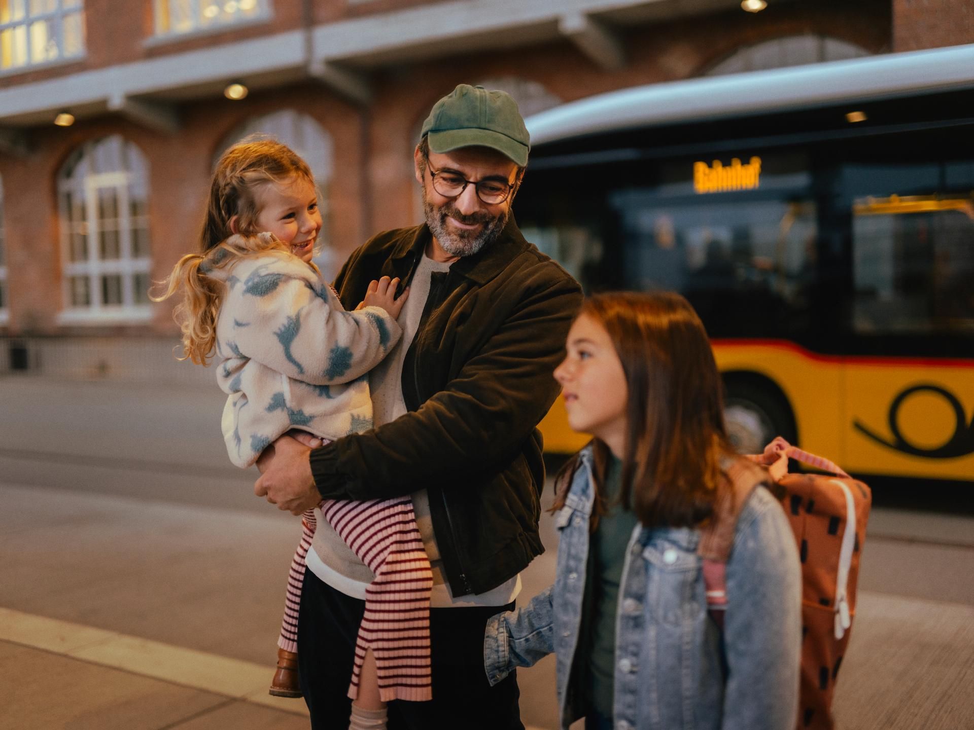 Three people are standing at a bus stop in front of a yellow bus. One person is holding a child, while another person with a backpack stands nearby. In the background, a brick building with illuminated windows is visible.
