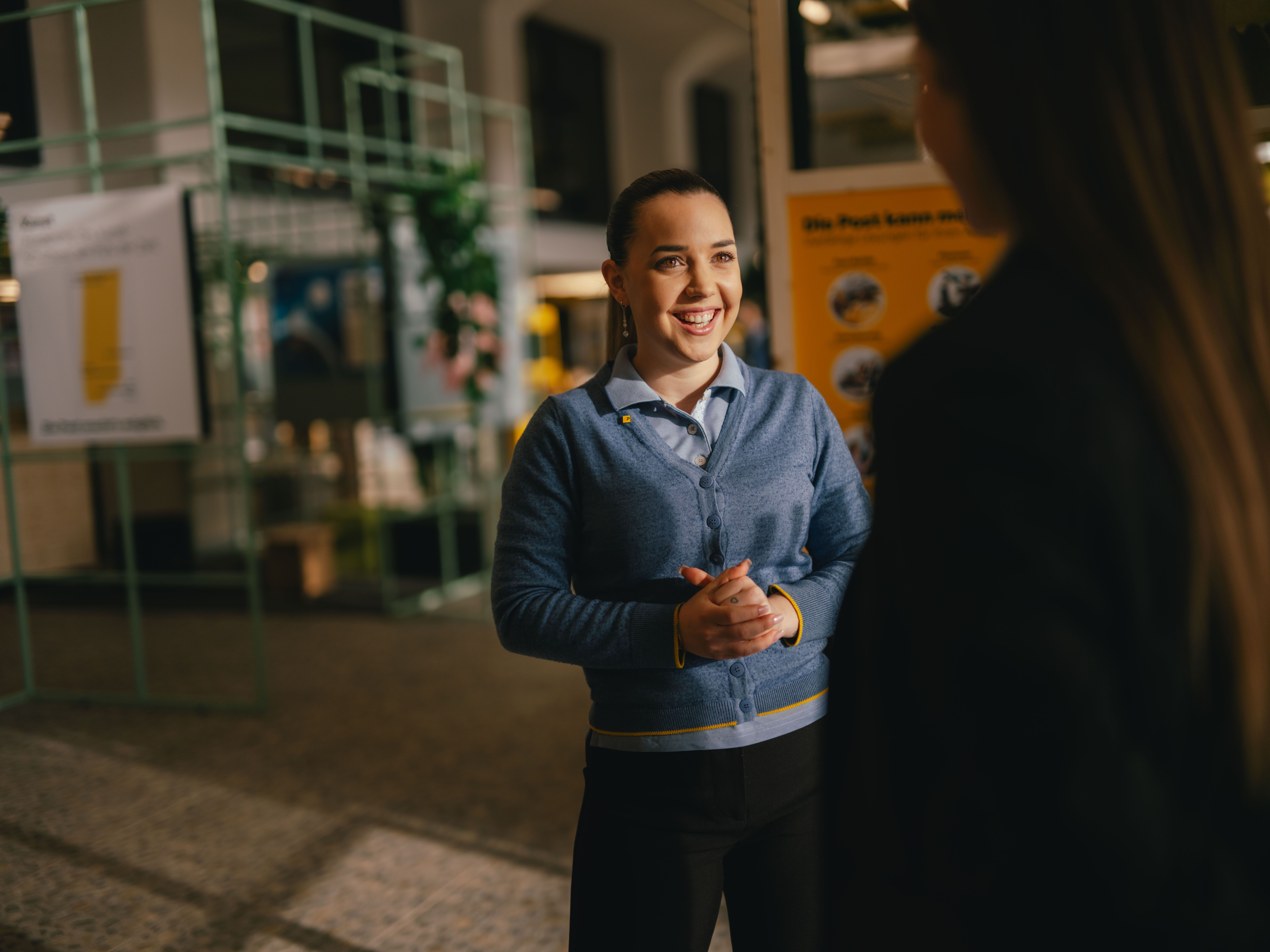 A person stands indoors and is engaged in conversation with another person in the foreground. The person is wearing a blue cardigan over a shirt and has hands clasped in front. In the background, informational displays and exhibition structures are visible.
